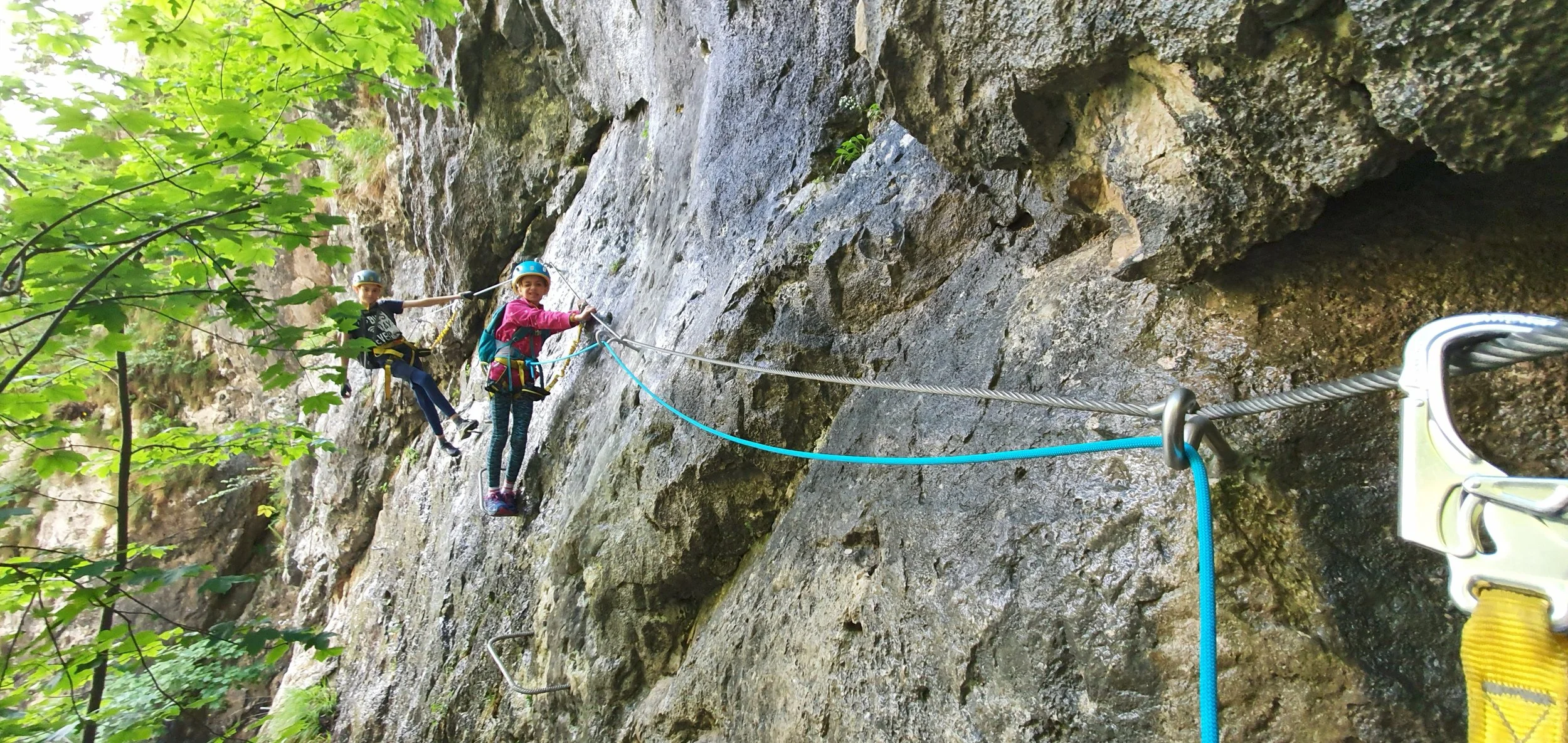 Two children rock climbing outdoors on a steep rocky cliff, wearing helmets and harnesses, connected by safety ropes, with green foliage in the foreground.