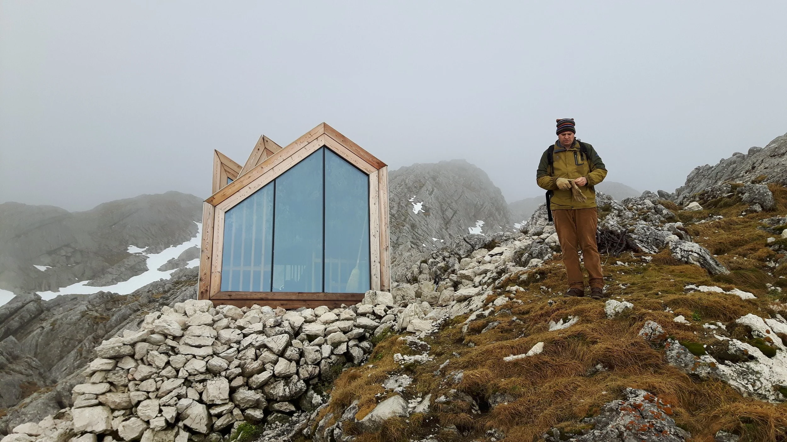 A person standing on rocky terrain near a modern, wooden and glass mountain hut with a foggy mountain backdrop.