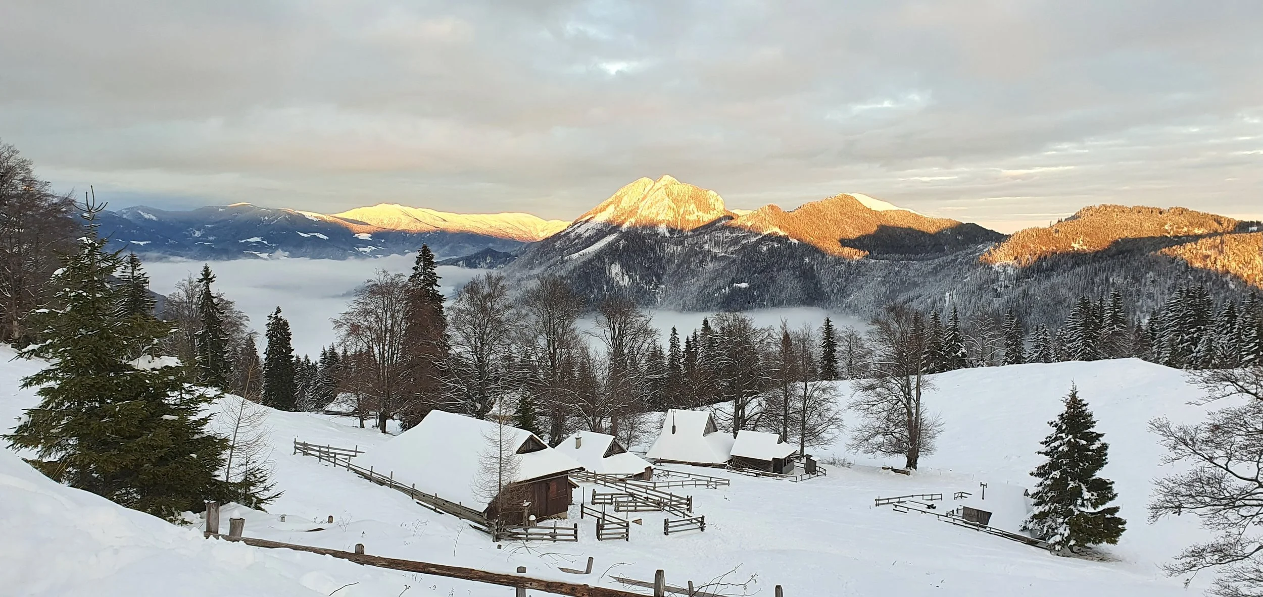 Snow-covered mountains and trees in a winter landscape with a small cabin and fence in the foreground, sunset illuminating the peaks.