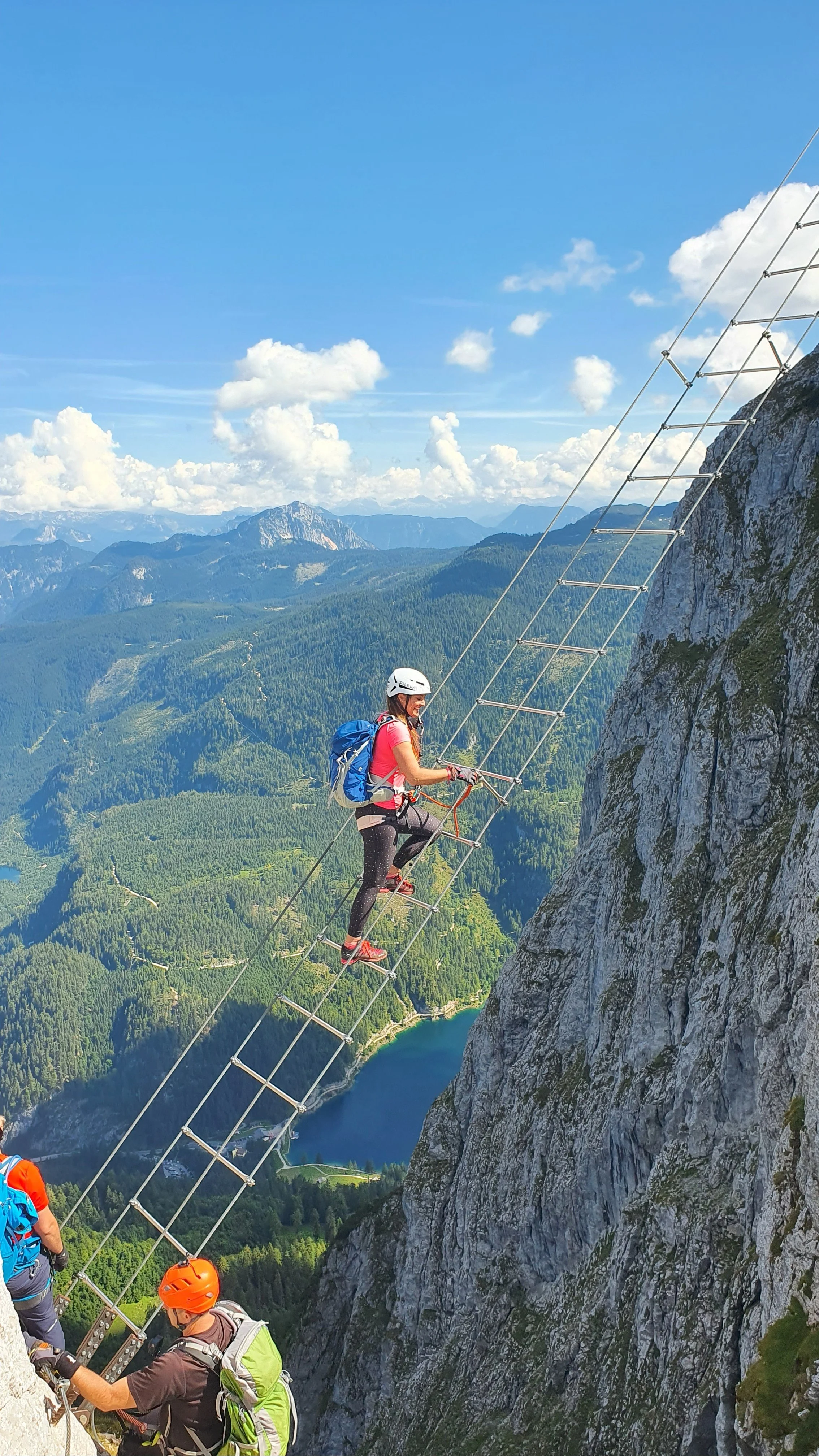 A woman wearing climbing gear, helmet, and backpack climbing a metal ladder on a mountain slope, with a scenic mountain landscape and lake in the background.