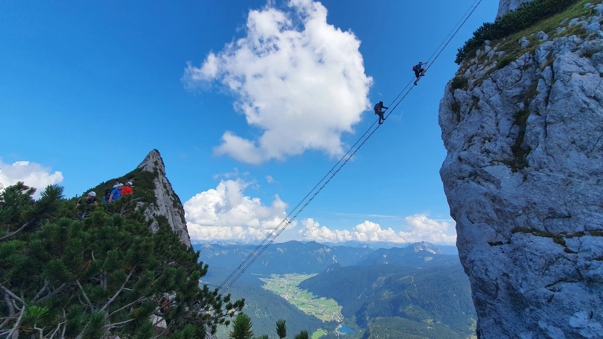 Group of hikers crossing a high suspension bridge between mountain cliffs with a scenic valley view below on a clear day.