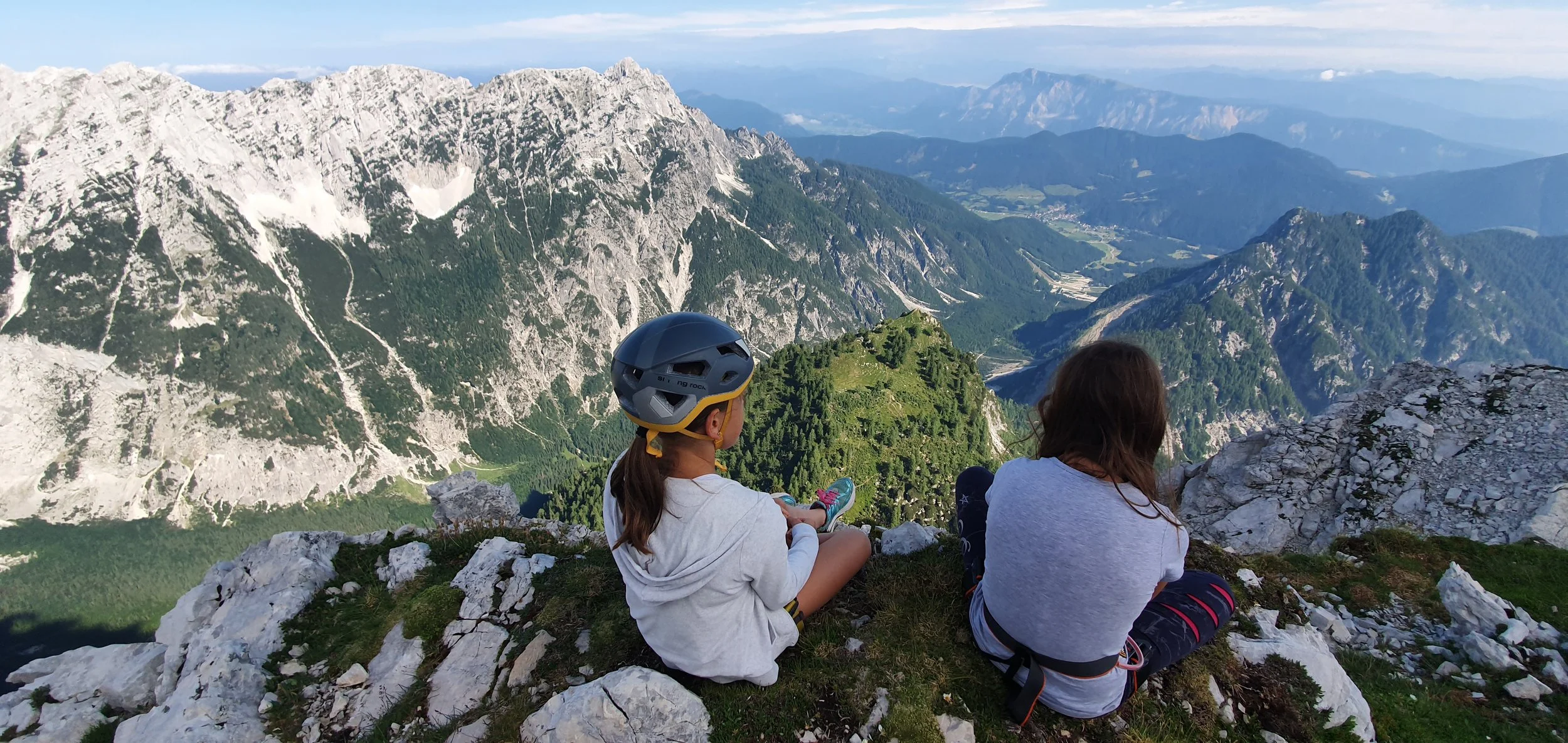 Two children sitting on a rocky mountain ledge, overlooking a scenic mountain landscape with peaks, forests, and valleys in the distance.