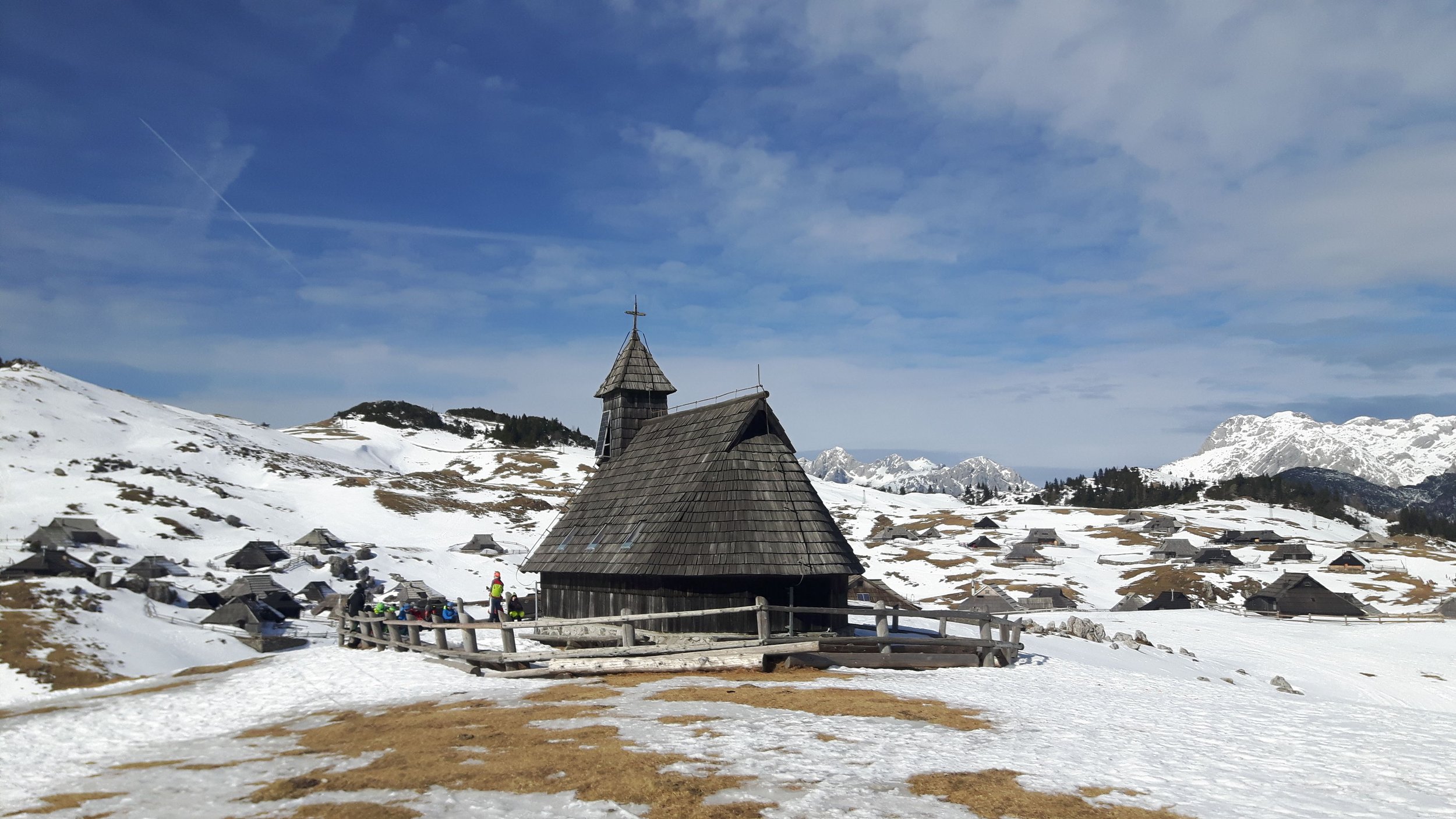 Snow-covered mountain landscape with a wooden church and scattered wooden houses, mountain peaks in the background, and a partly cloudy sky.