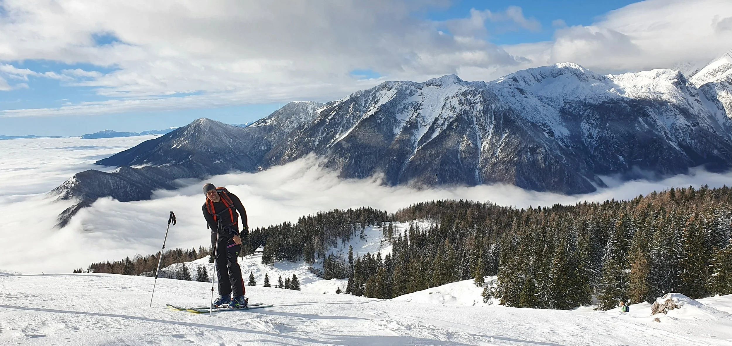 A person skiing on snowy mountain terrain with a backdrop of snow-capped mountains, clouds, and a forest of pine trees.