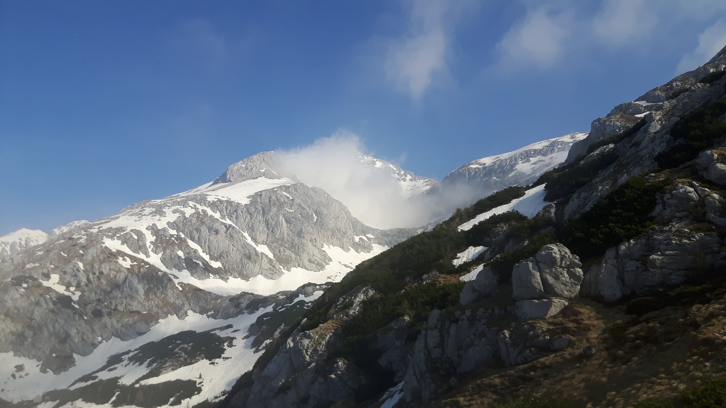 Snow-capped mountains with rocky slopes and patches of greenery beneath a blue sky with some clouds.