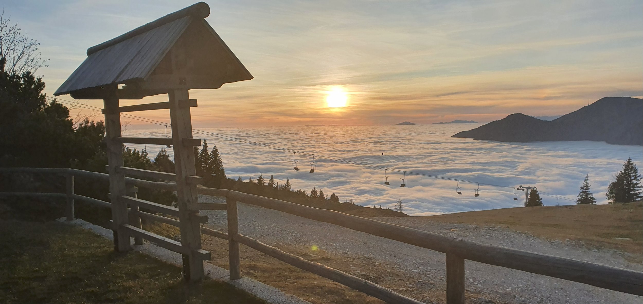 Sunset over a mountain landscape with clouds below, ski lift chairs hanging above, and a wooden hut in the foreground.