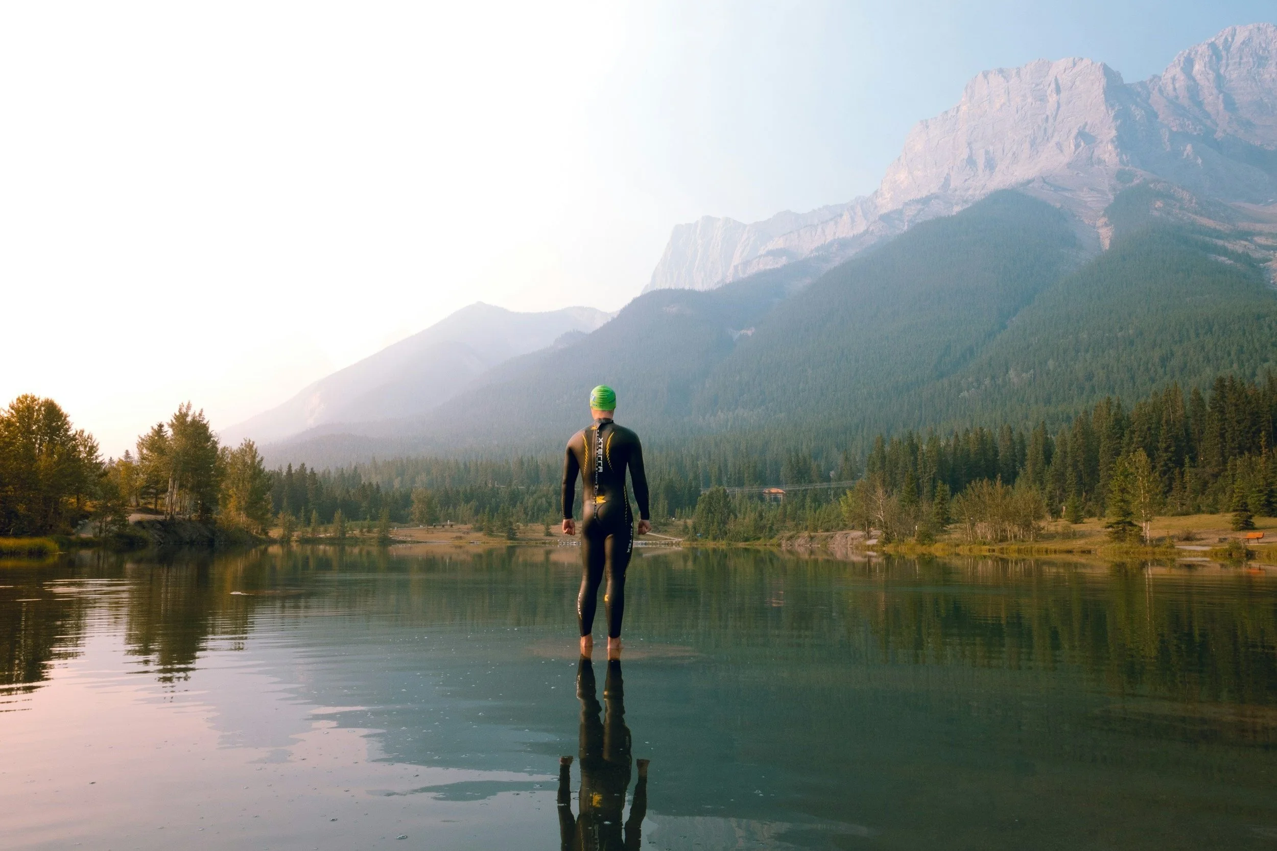 Swimmer standing on the edge of a calm lake with mountains and forest in the background.