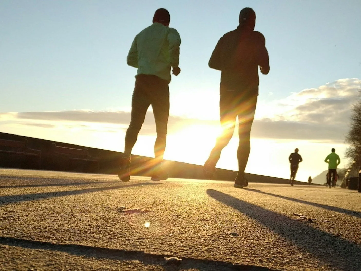 People jogging on a paved path during sunrise with the sun shining in the background, casting long shadows.