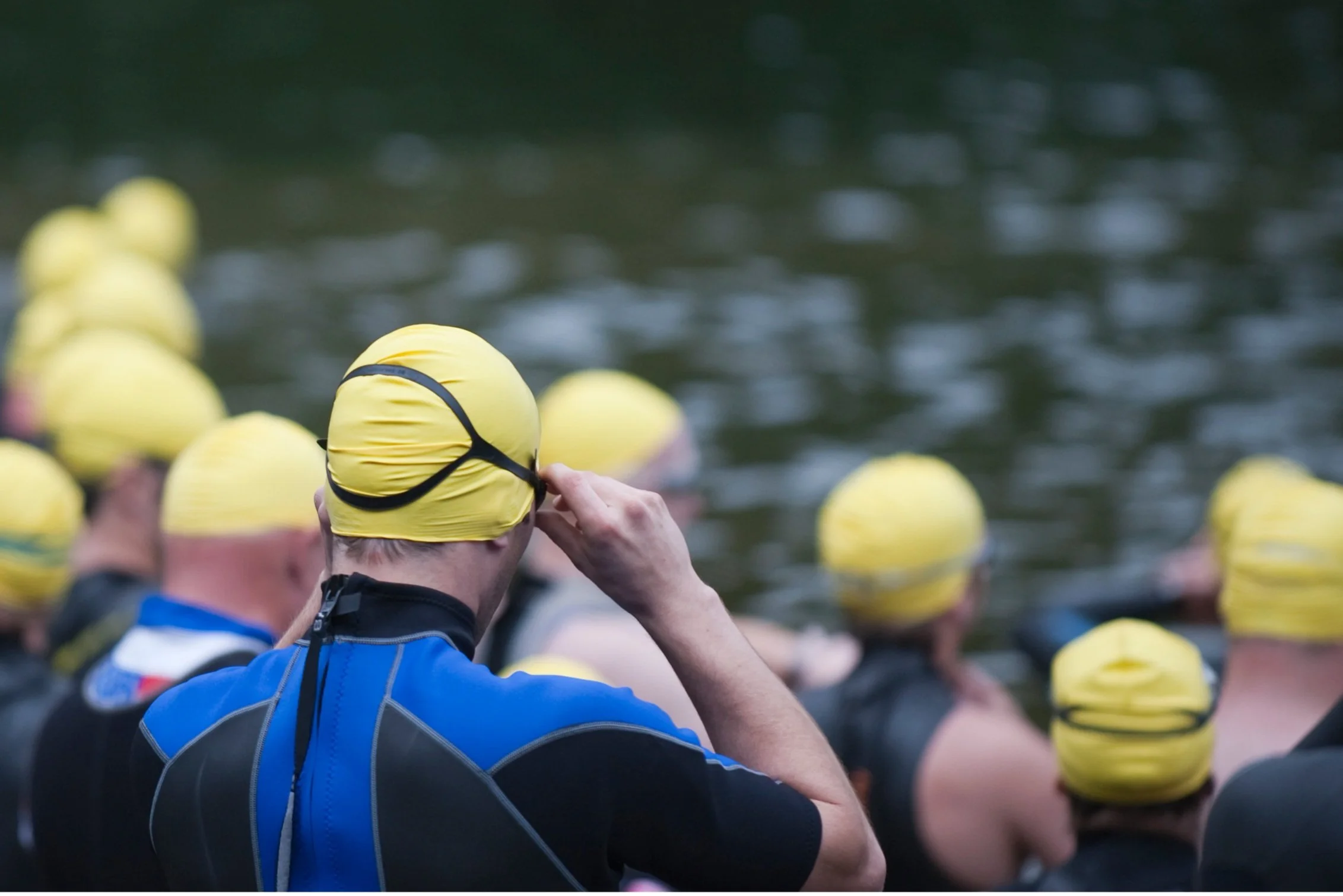 Swimmers in black wetsuits and yellow swim caps preparing for a race by the water.