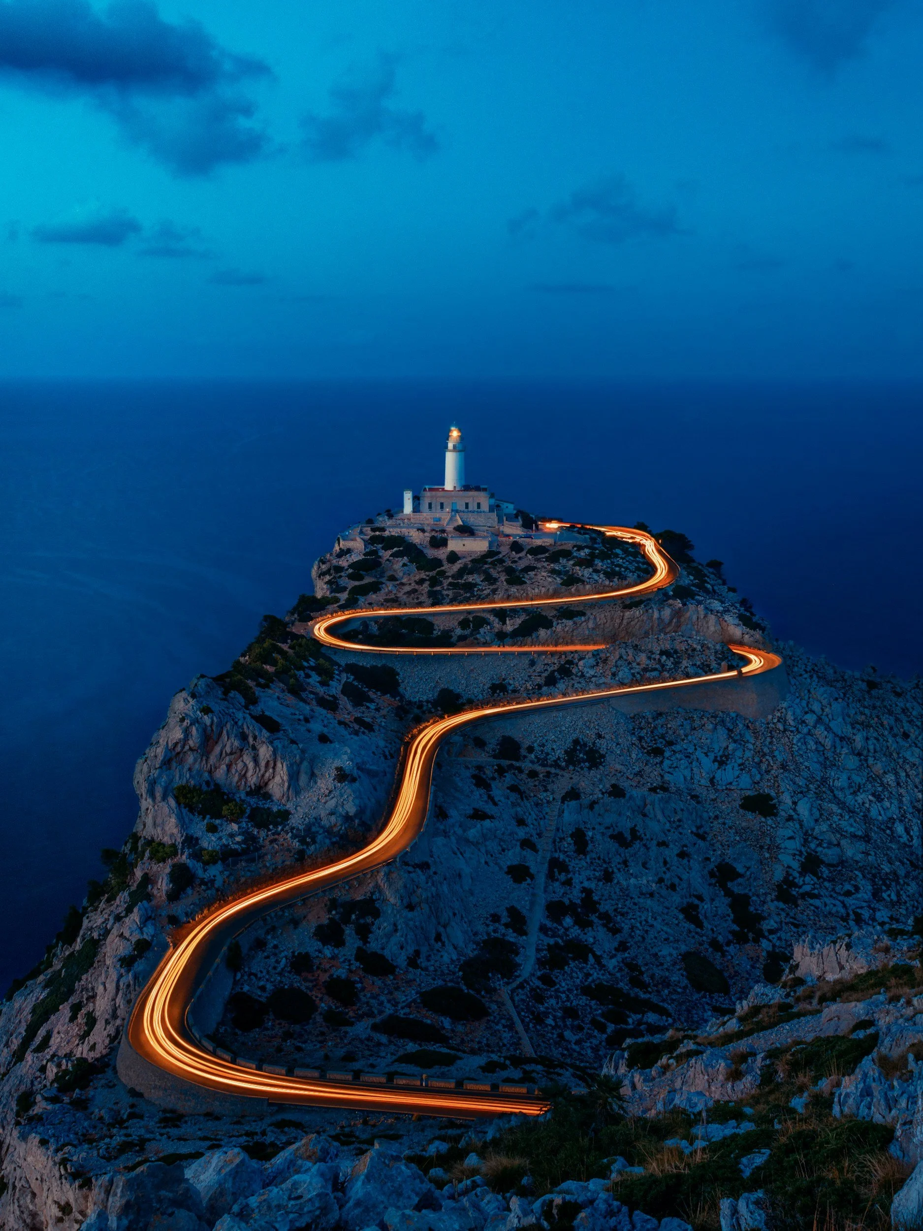 Nighttime view of a winding mountain road leading to a lighthouse on a cliff, with light trails from vehicles and a cloudy sky overhead.