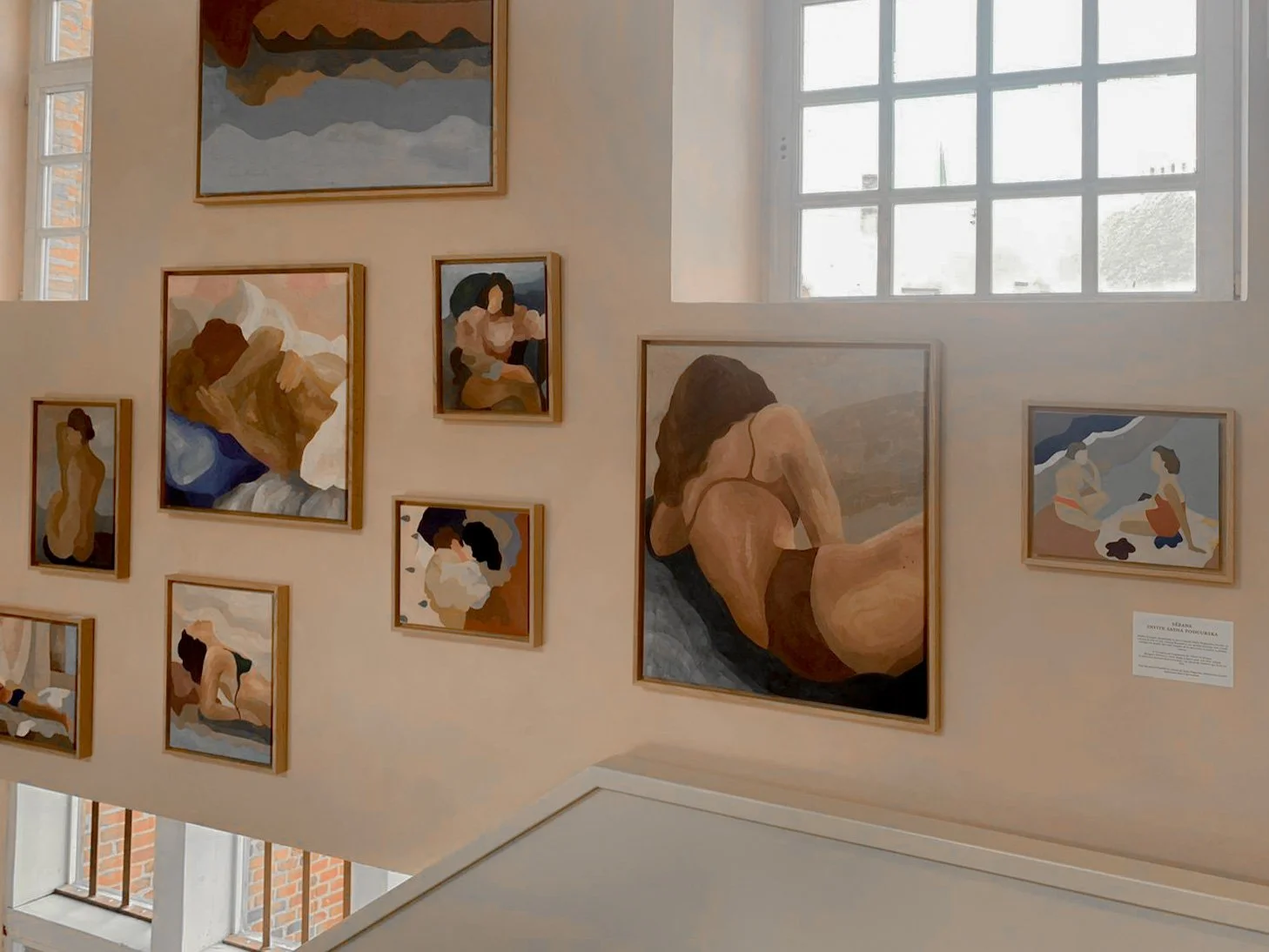 Une galerie d'art avec plusieurs peintures modernes représentant des femmes dans diverses poses, accrochées à un mur clair près de fenêtres laissant entrer la lumière naturelle.
