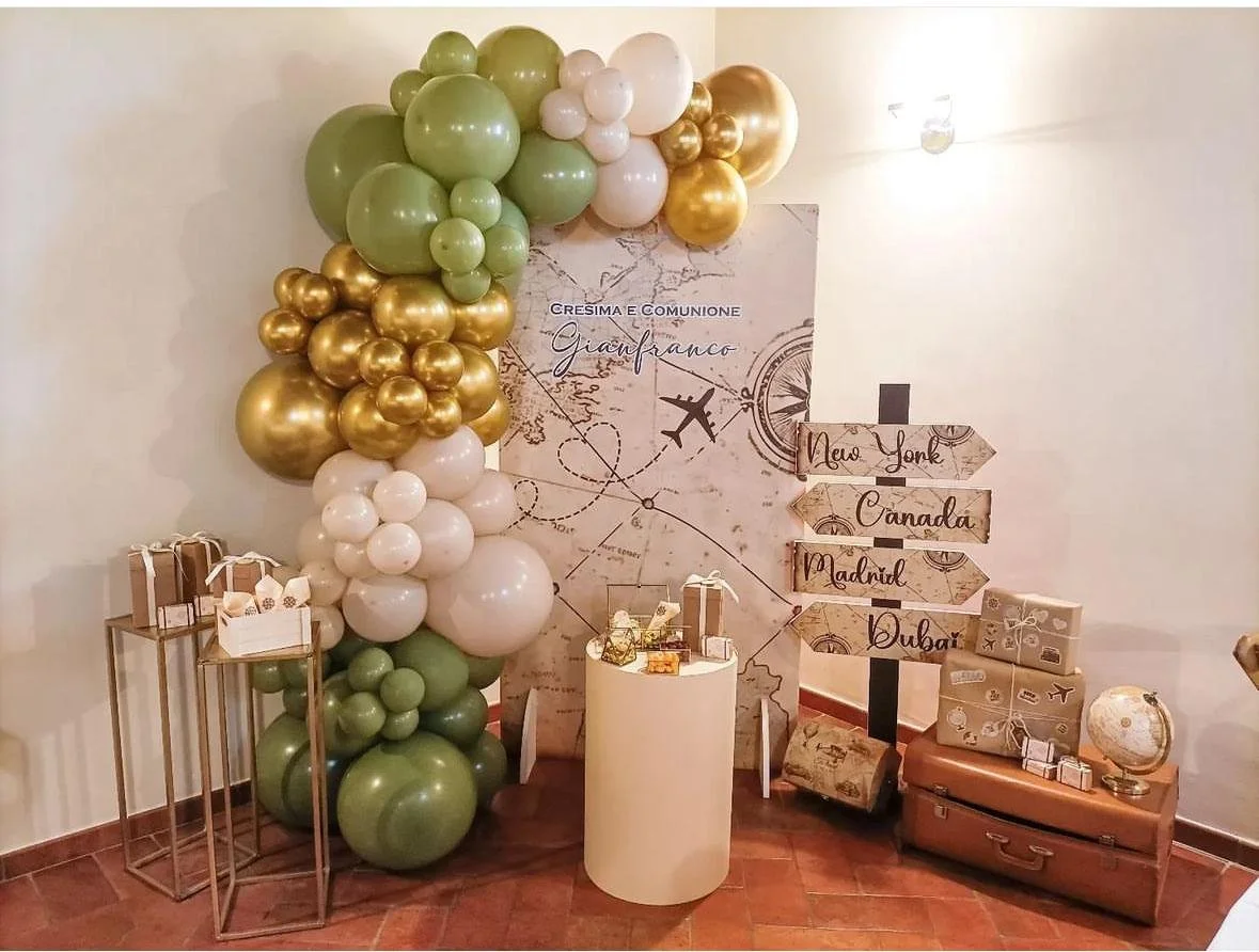 Decorazione per comunione con palloncini verdi, bianchi e dorati, con cartelli che indicano città come New York, Canada, Madrid e Dubai, e scatole regalo dov'è posizionato anche un globo terrestre.
