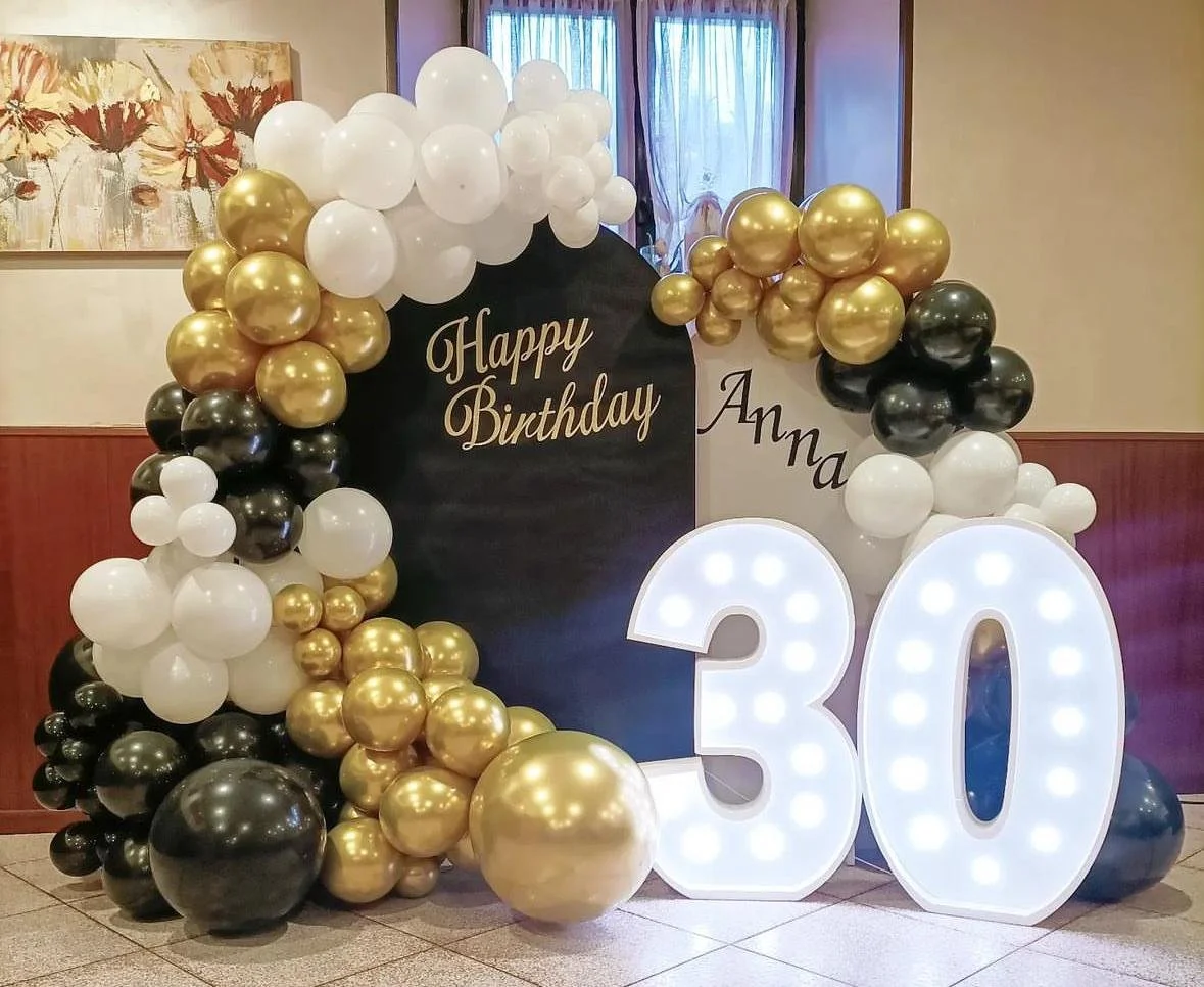 Decorazione di compleanno con palloncini bianchi, neri e oro, grandi numeri luminosi '30' e messaggio 'Happy Birthday Anna' su uno sfondo nero