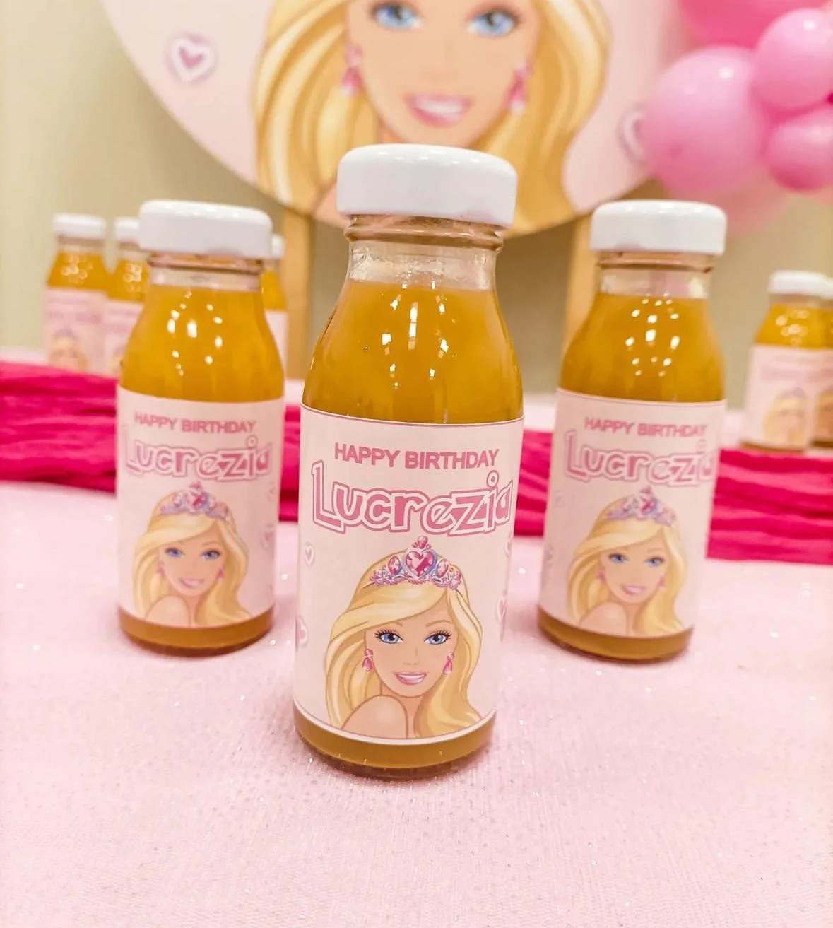 Mini bottiglie di succo con etichetta personalizzata per il compleanno di Lucrezia, decorazioni rosa e una grande illustrazione di una ragazza con capelli biondi e coroncina.