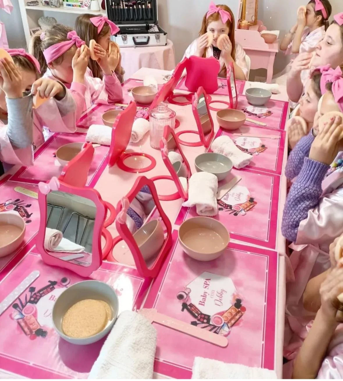 Gruppo di bambine con asciugamano di stoffa bianco e visiera di cartone rosa, che partecipano a un'attività di trucco e bellezza in un ambiente rosa, con tavolo decorato con set di trucco e specchi.