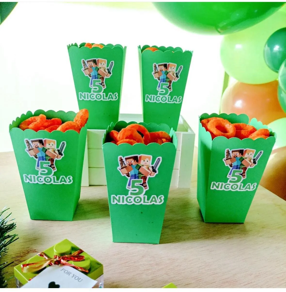 Contenitori verdi a forma di popcorn, decorati con un'etichetta di Minecraft con due personaggi e il numero 5, contenenti snack arancioni, posti su un tavolo per una festa di compleanno.