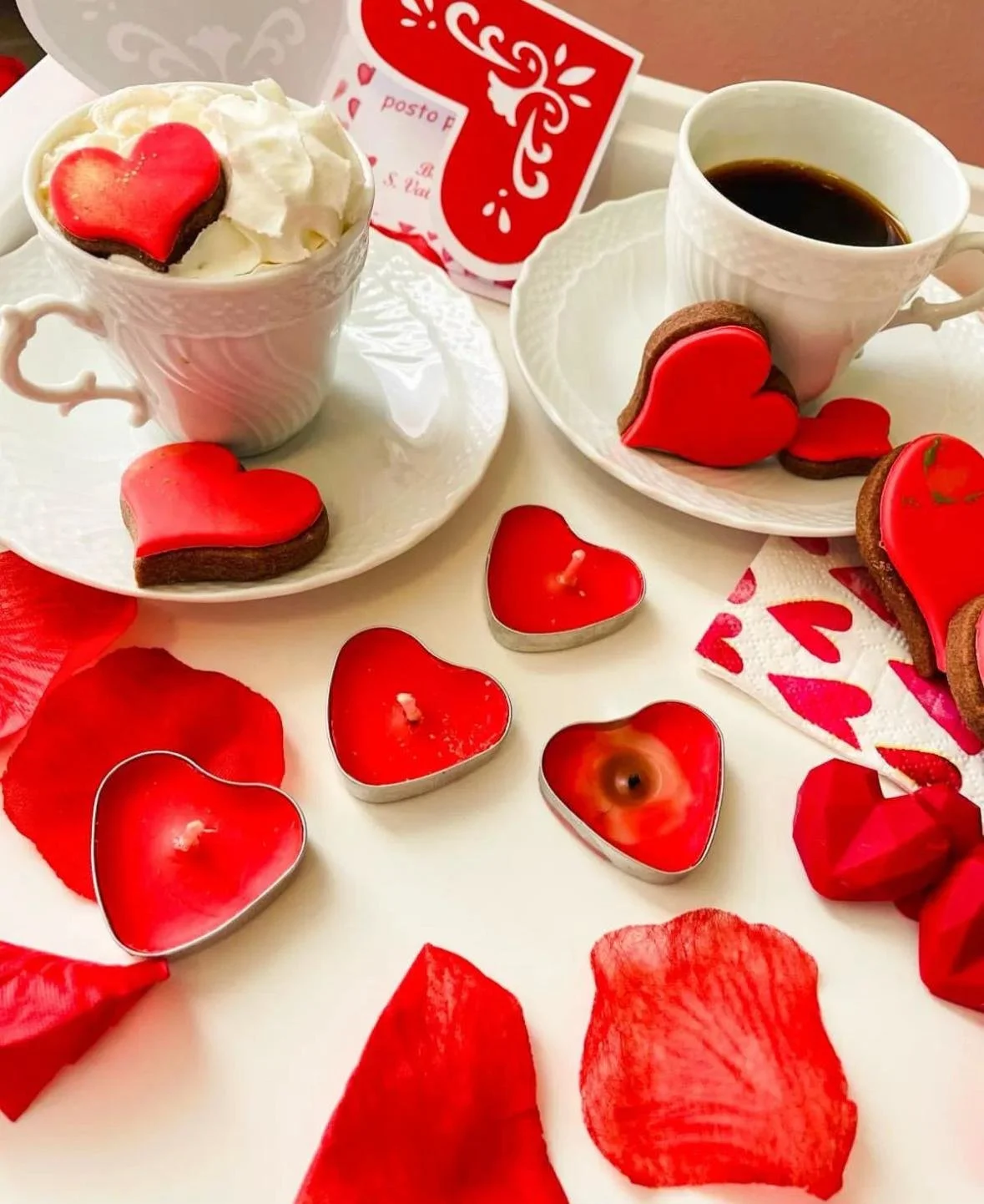 Tazze di caffè con panna e biscotti a forma di cuore decorati con glassa rossa, oltre a candele a forma di cuore, petali di rosa e decorazioni a tema san Valentino.