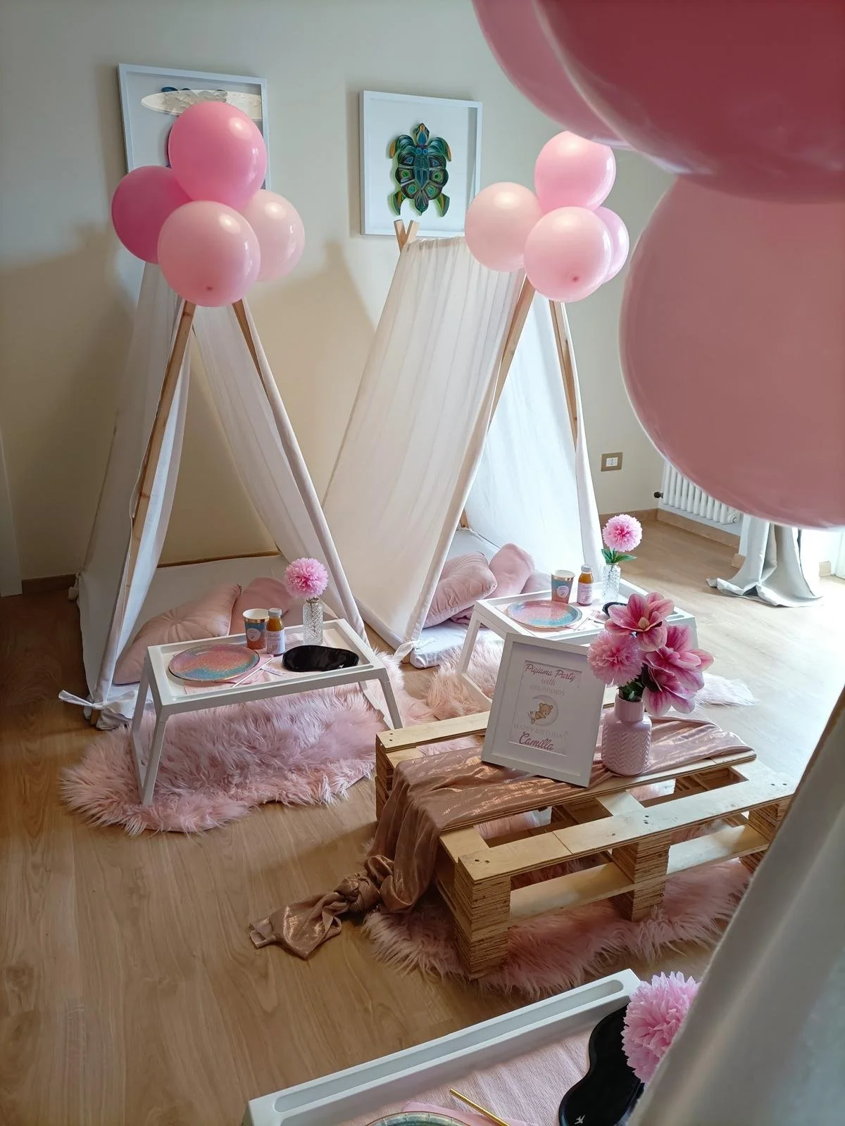 Decorazione per festa di bambina con tende bianche, palloncini rosa, fiori rosa in vasi, tavolini bianchi con piatti colorati, un cartellino con scritto 'Pinkaline Party con Camilla', tappeti rosa e un drappeggio di tessuto rosa