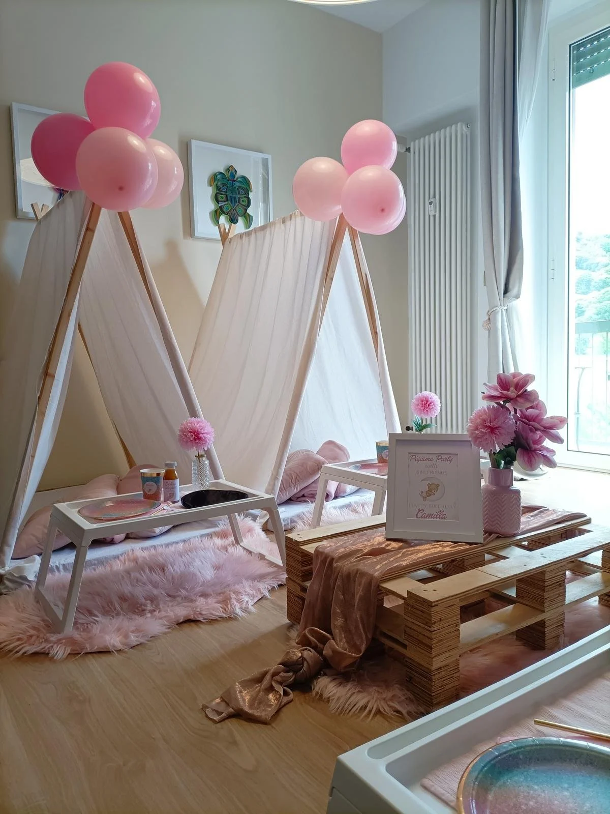 Decorazione per festa con tende a cielo, palloncini rosa, fiori rosa, tavolino e poster con scritte in italiano.