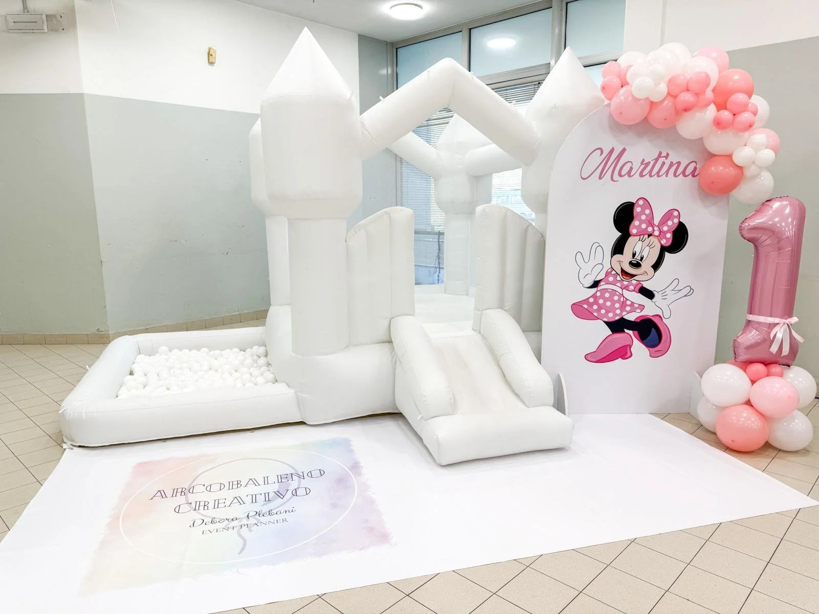 Decorazione di compleanno con castello gonfiabile bianco, un grande cartellone con Minnie Mouse, palloncini rosa e bianchi con il numero uno, e un tappeto con scritte. Il cartellone dice 'Martina' e mostra Minnie Mouse in posa.