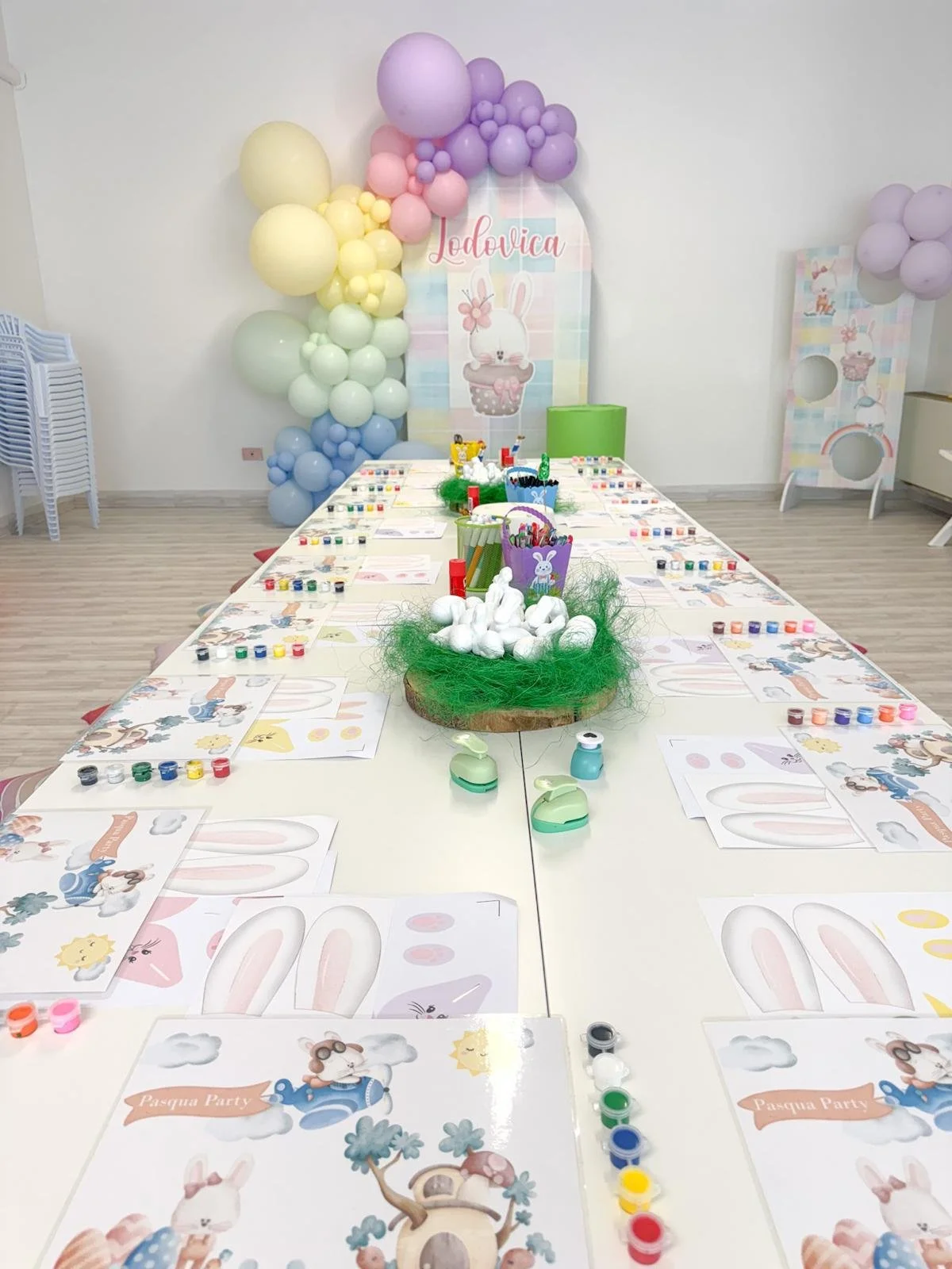 Tavolo decorato per una festa di Pasqua con disegni di conigli, uova e altri simboli pasquali, con materiali per attività artistiche e colorate, tra palloncini pastello rosa, gialli, verdi e blu e uno sfondo con un coniglietto e il nome Ludovica.