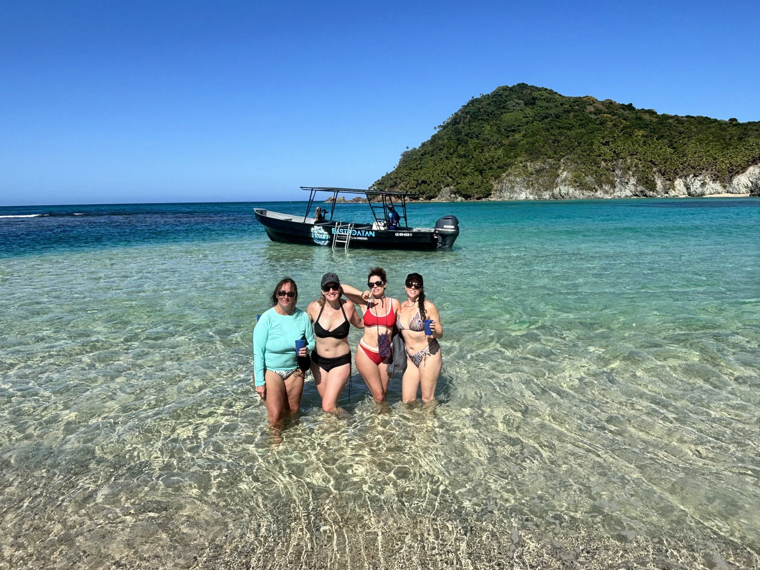 Four women standing in shallow, clear ocean water near a boat and a green island in the background on a sunny day.