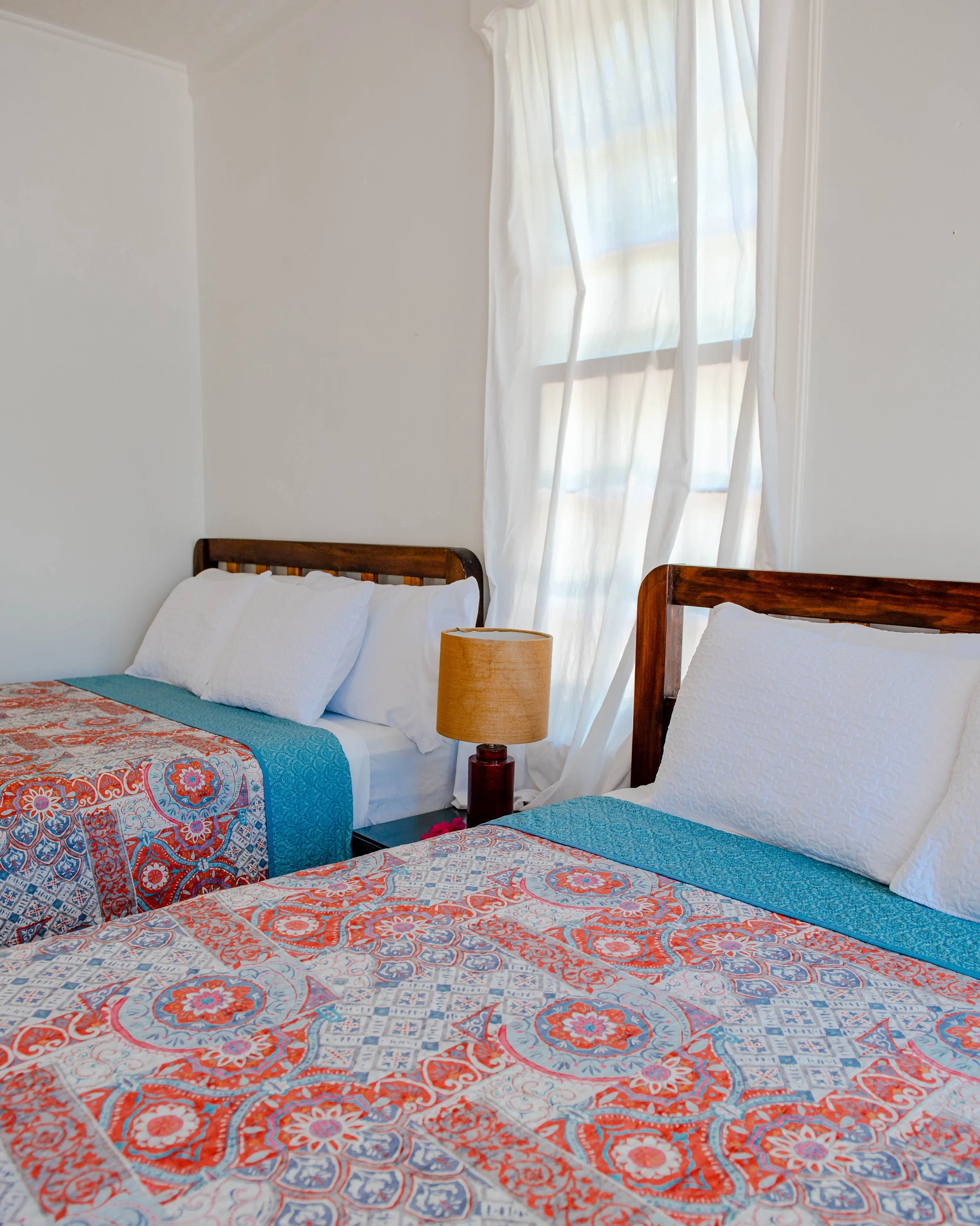 Two beds with colorful patterned bedspreads, white pillows, a table lamp, and a window with white curtains in a bright bedroom.