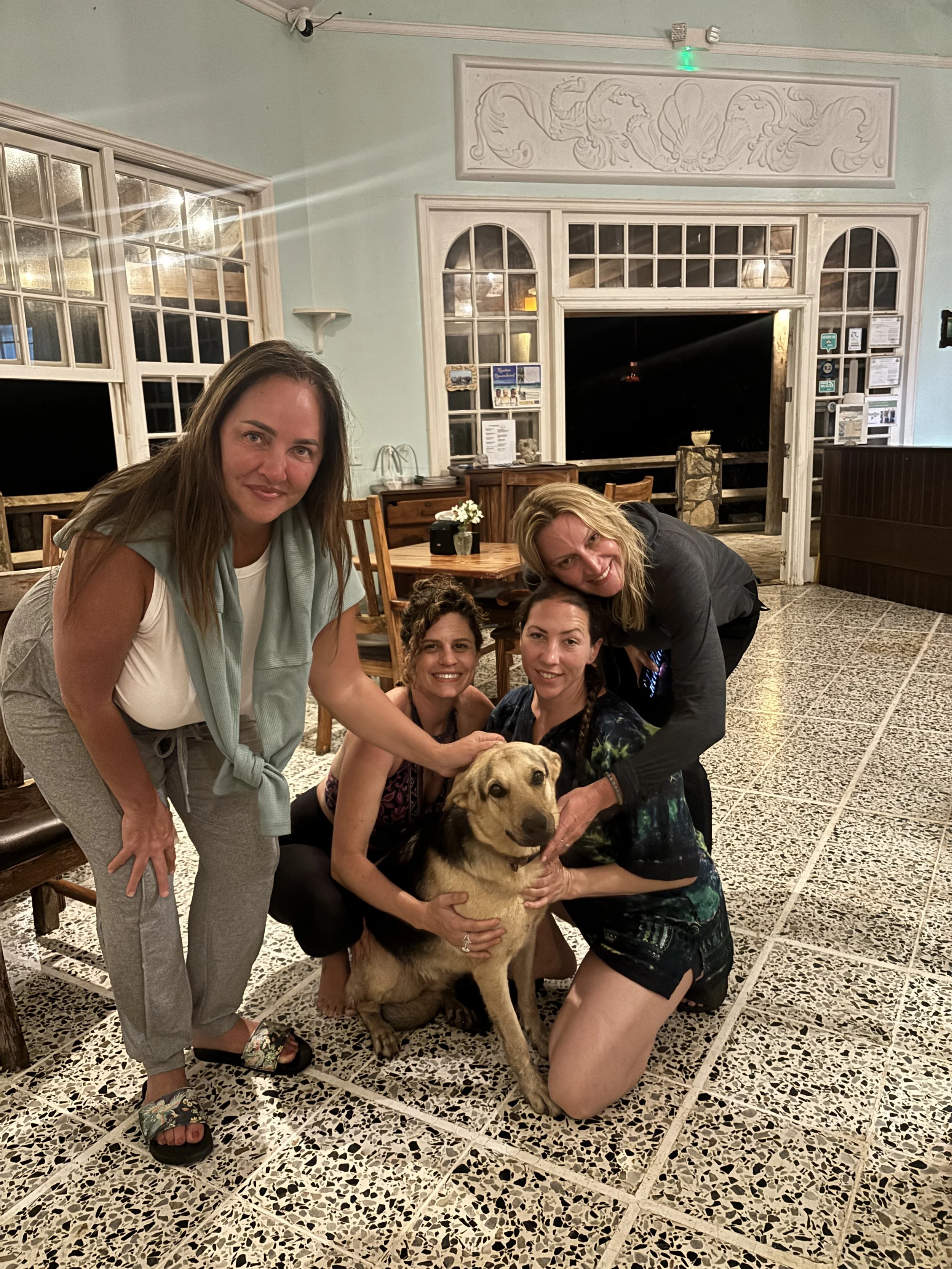 Four women and a dog posing inside a restaurant at night. The women are smiling and holding the dog, which is sitting on the tiled floor. The background features large windows, wooden furniture, and decorative wall elements.