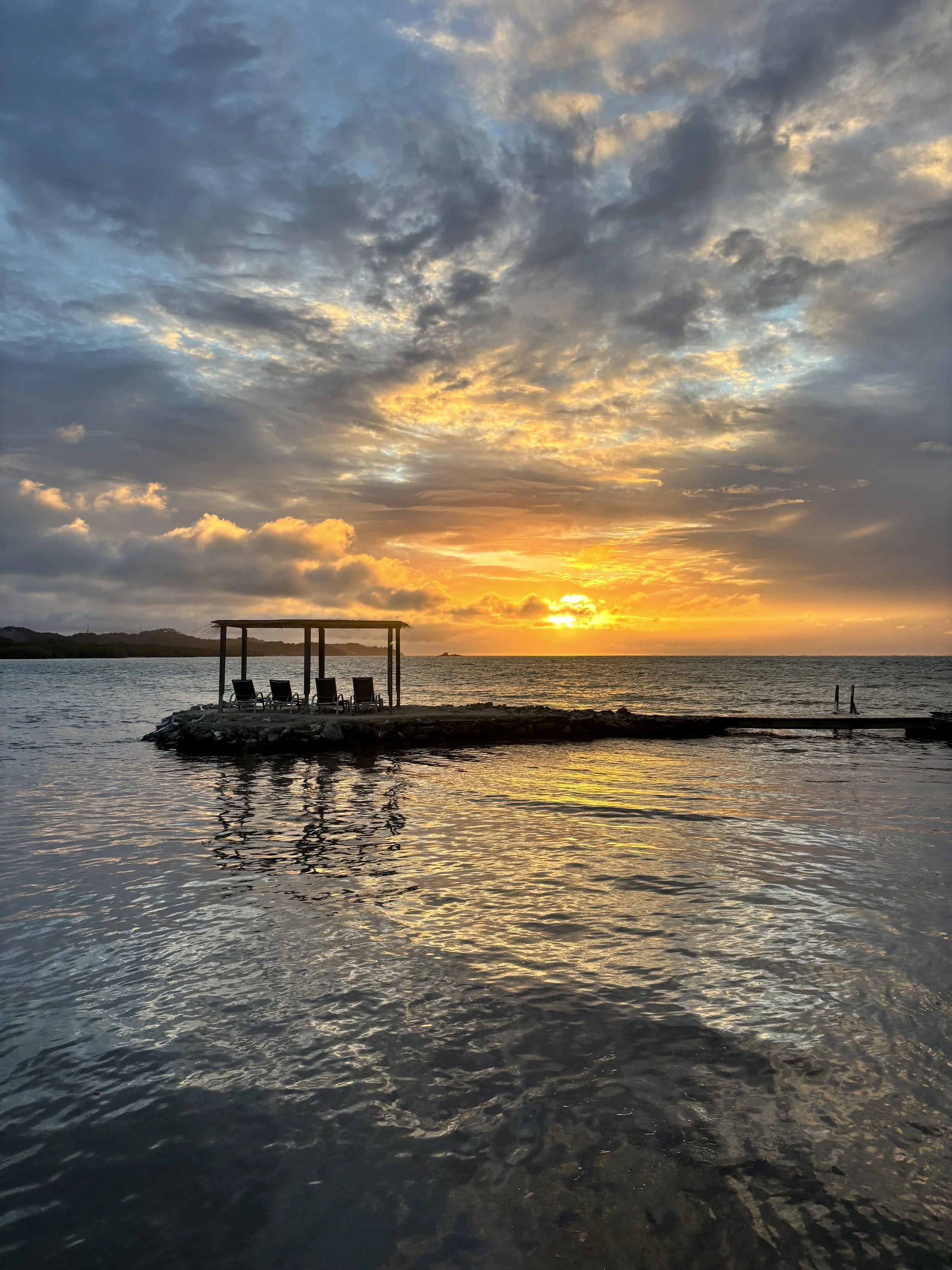 Calm ocean water reflecting a vibrant sunset sky with clouds, a small pier with six chairs under a canopy, and surrounding landscape.