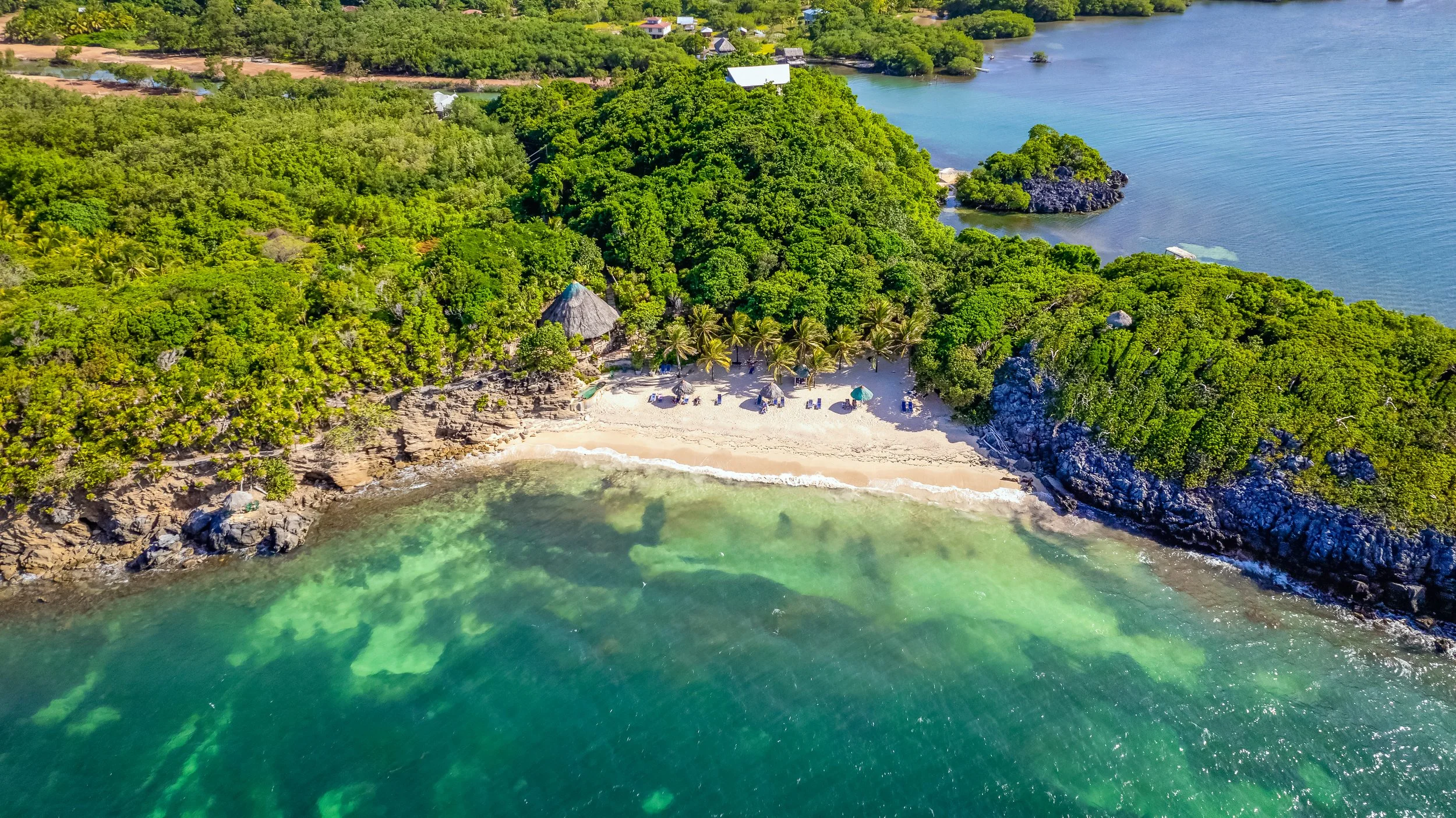 Aerial view of a tropical beach with clear water, sandy shoreline, lush green trees, and small cottages with thatched roofs.