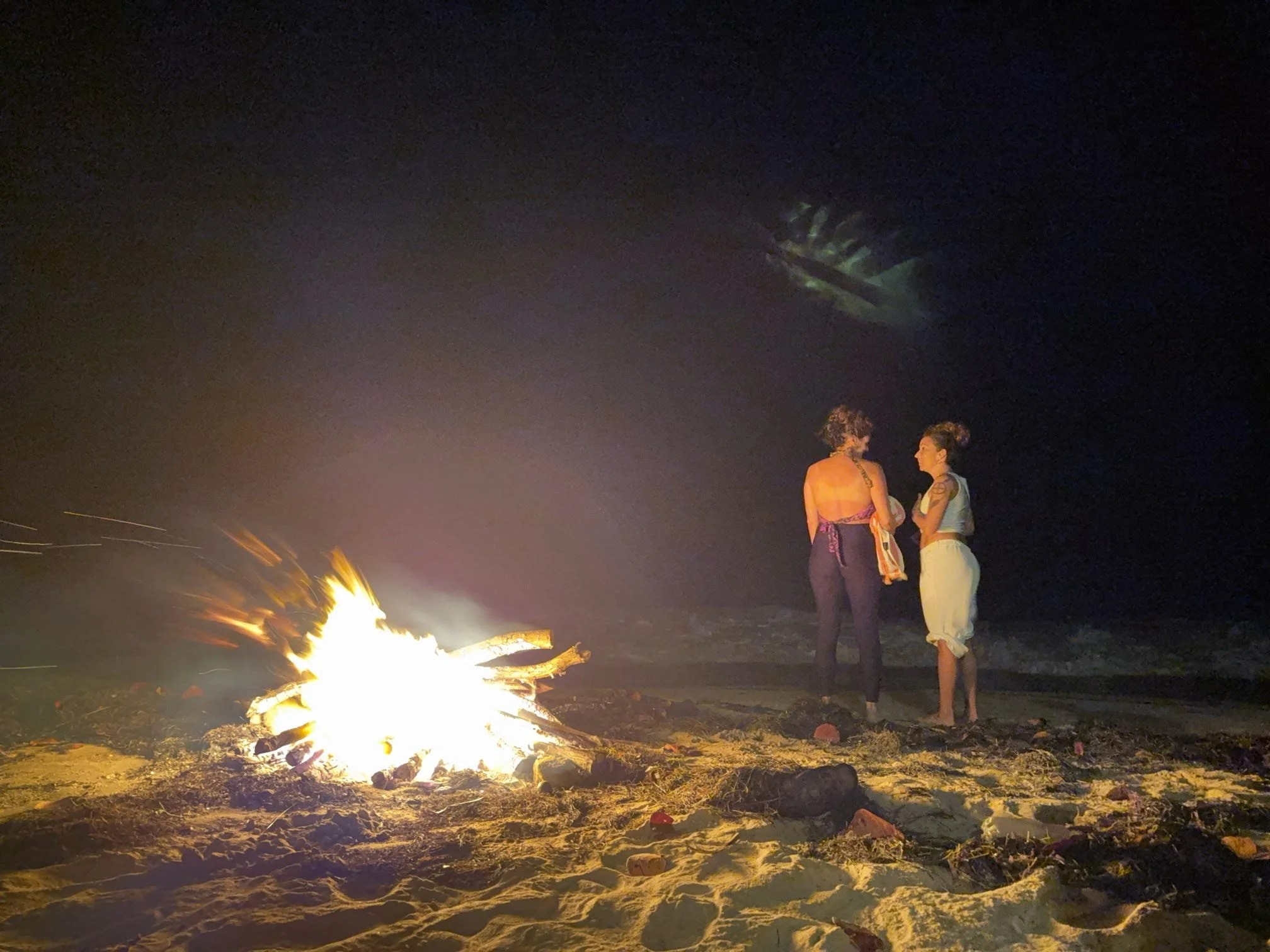 Two women standing on the beach at night near a campfire, facing each other and talking.