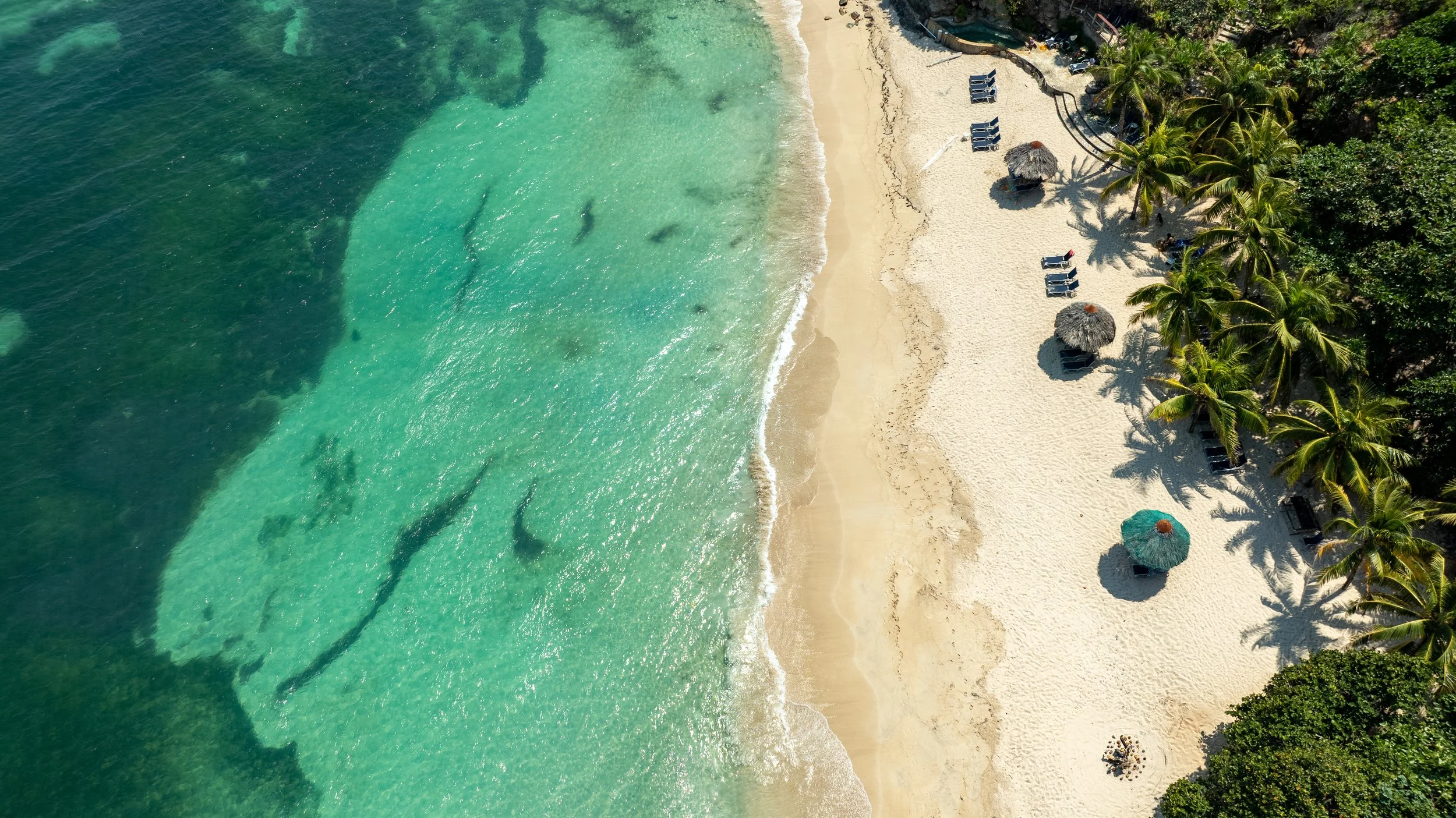 Aerial view of a tropical beach with clear turquoise water, white sandy shore, palm trees, and beach chairs with umbrellas.