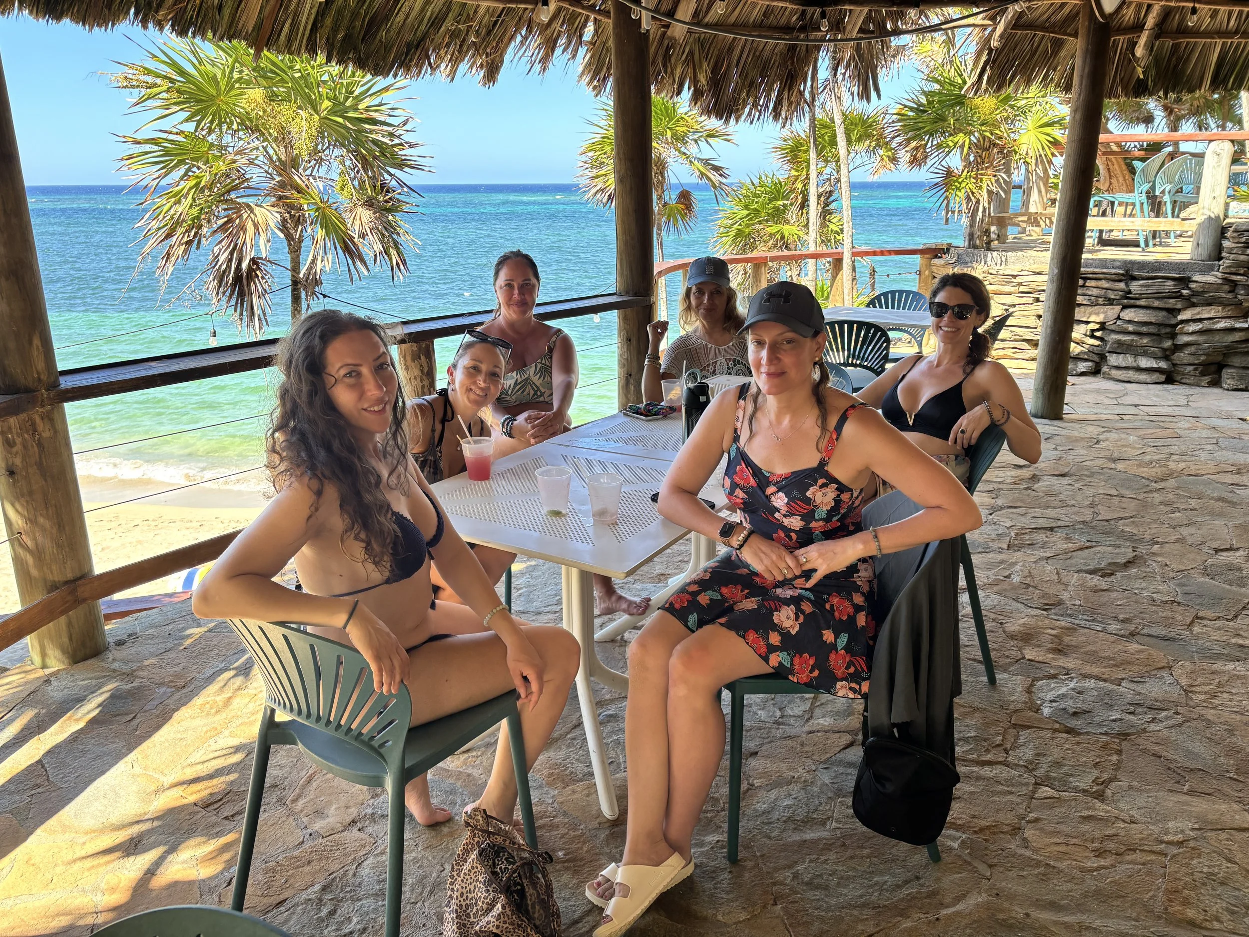 Six women sitting around a table at a beachside restaurant with a view of the ocean, palm trees, and a thatched roof overhead.