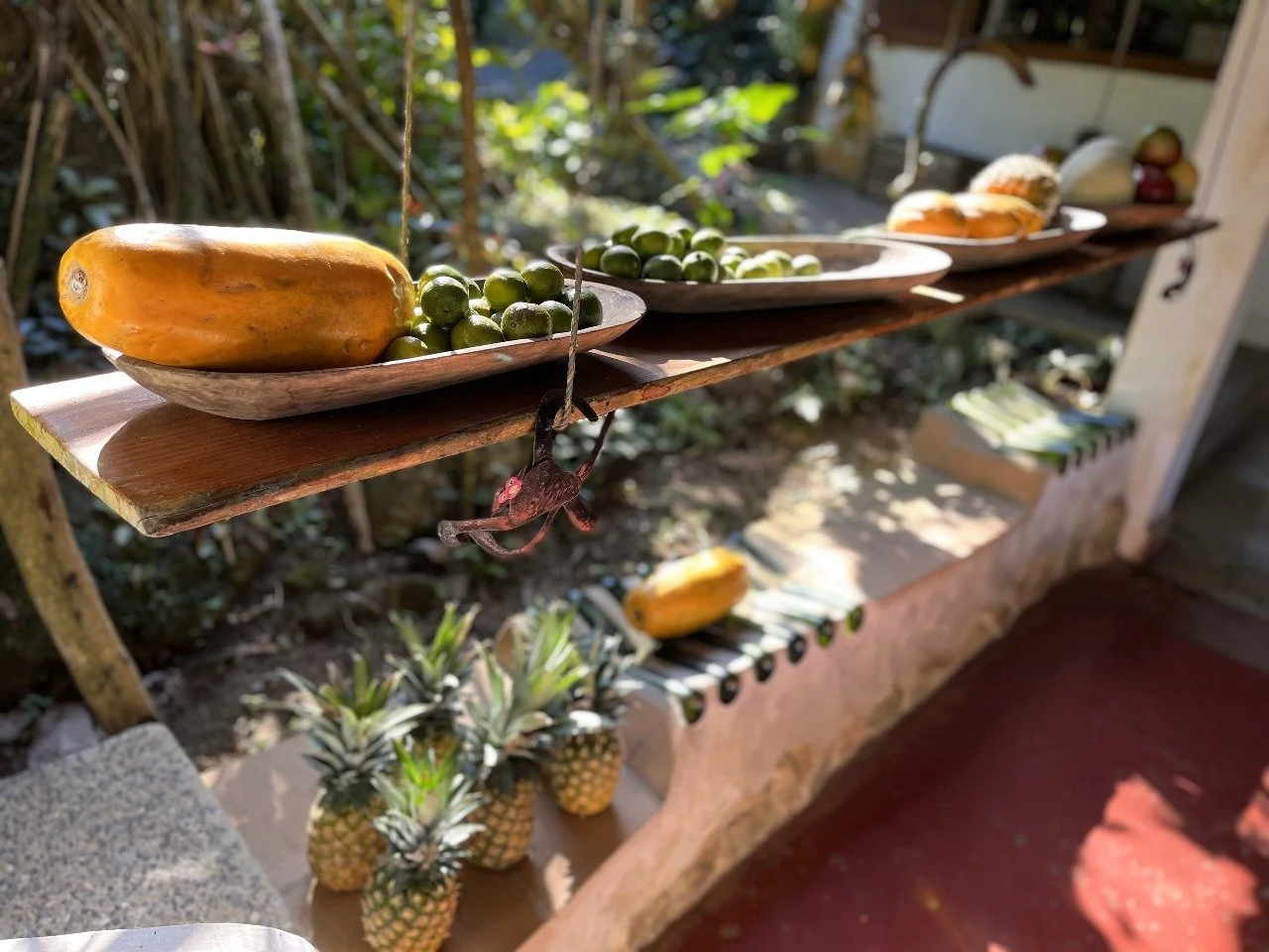 Outdoor display of fresh tropical fruits including papayas, green grapes, pineapples, and other fruits on wooden shelves.