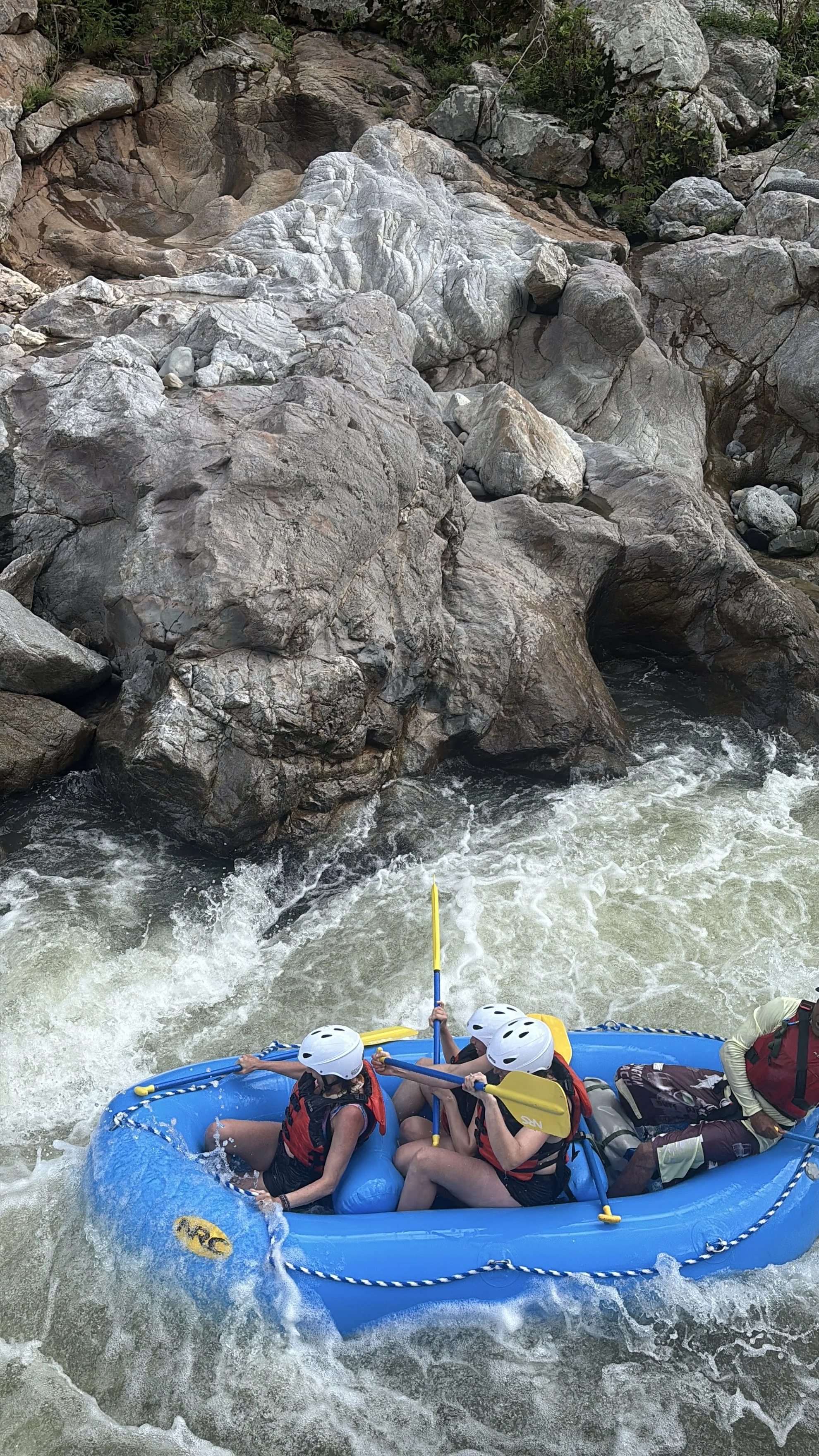 Four people in a blue inflatable raft white-water rafting on a river with rocks and rapids, wearing helmets and life jackets.