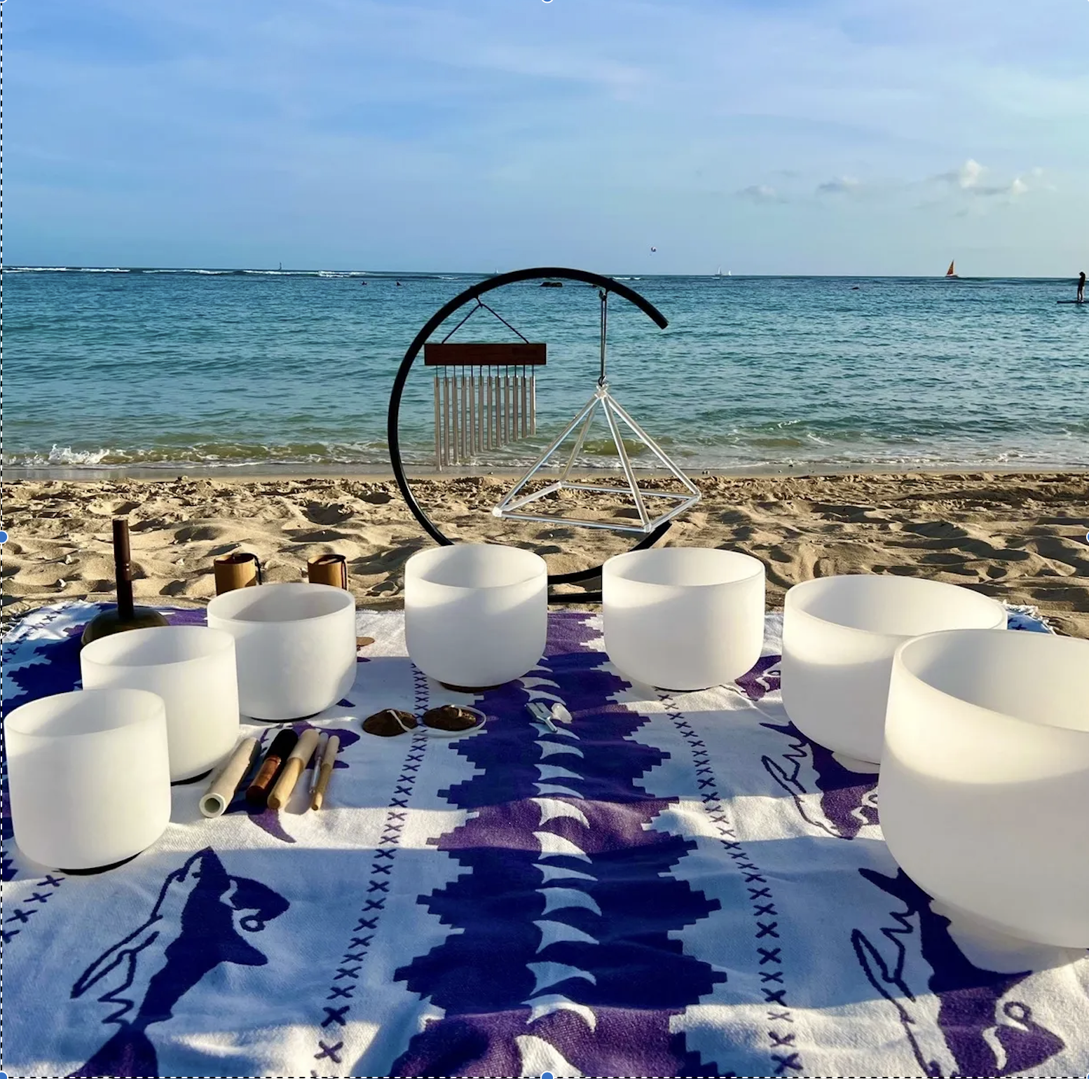 A beachside setup with singing bowls, mallets, and a decorative cloth, overlooking the ocean with a wind chime and geometric decorative piece in the background.