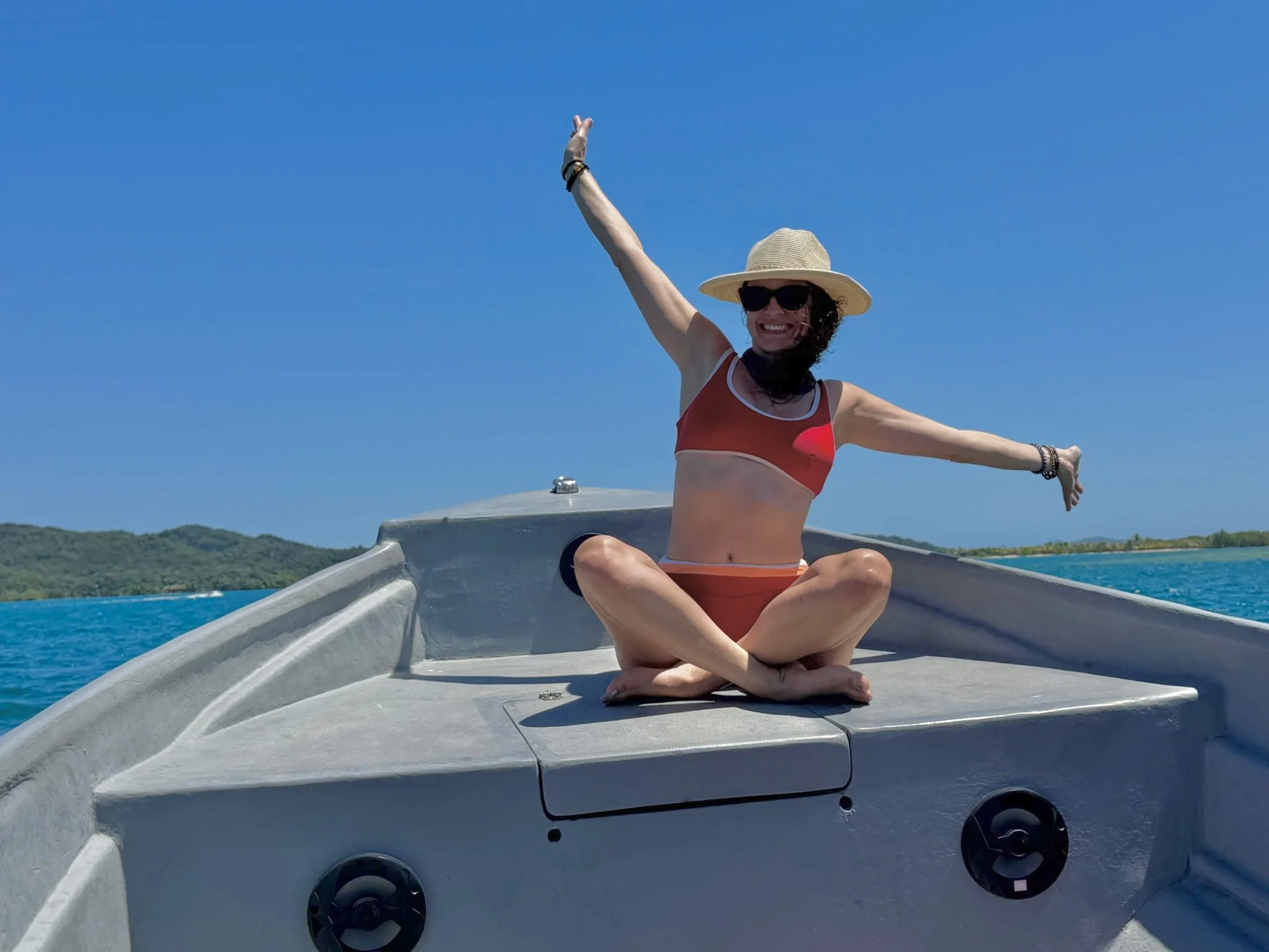 A woman sitting cross-legged on the top of a boat, wearing a red swimsuit, a straw hat, and sunglasses, smiling and raising her arms in the air, with clear blue sky and green land in the background.