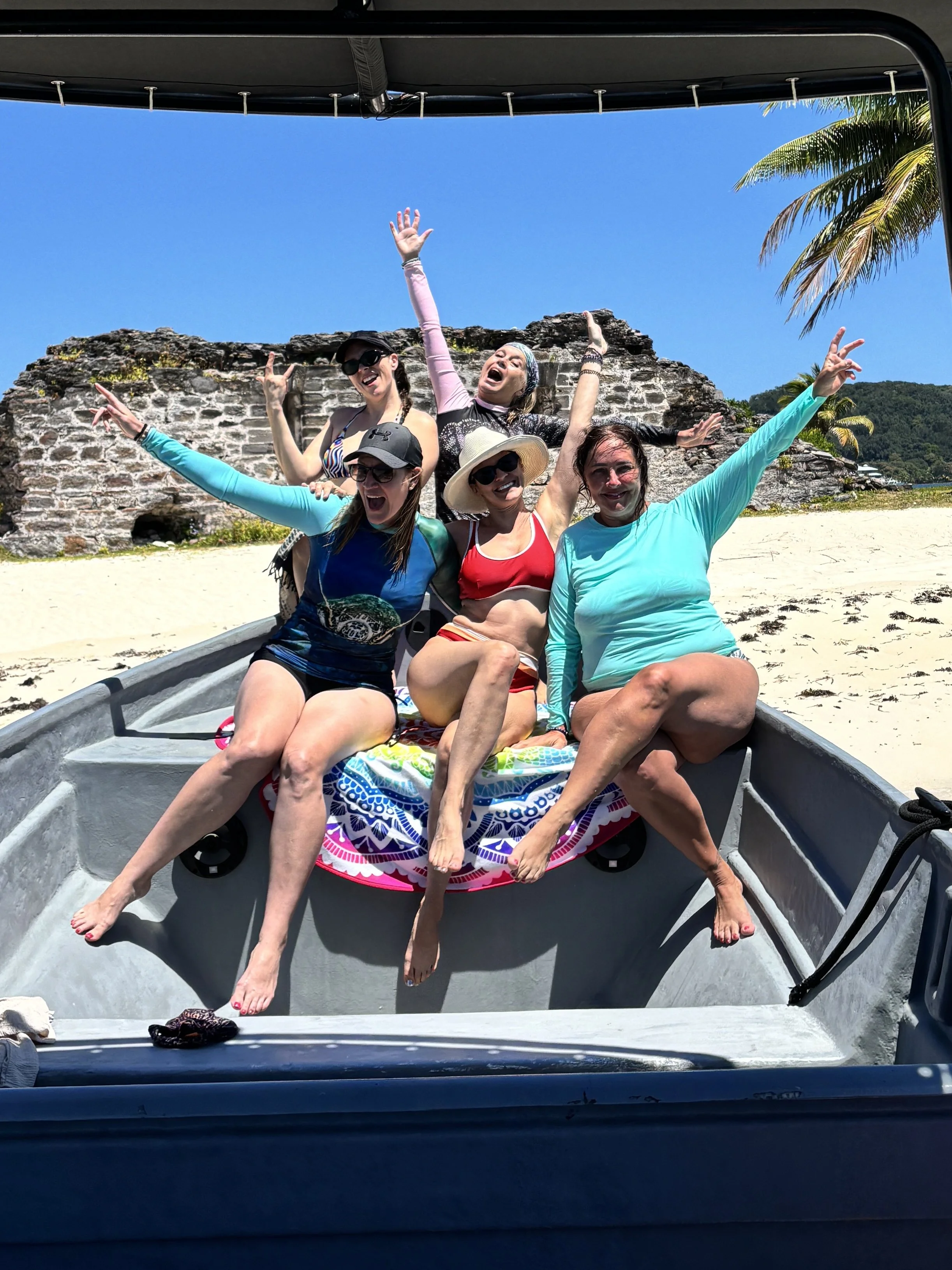 Five women sitting in the front of a boat on a beach, with a stone structure and palm trees in the background. They are wearing swimsuits and summer hats, smiling, laughing, and waving in the sunshine.