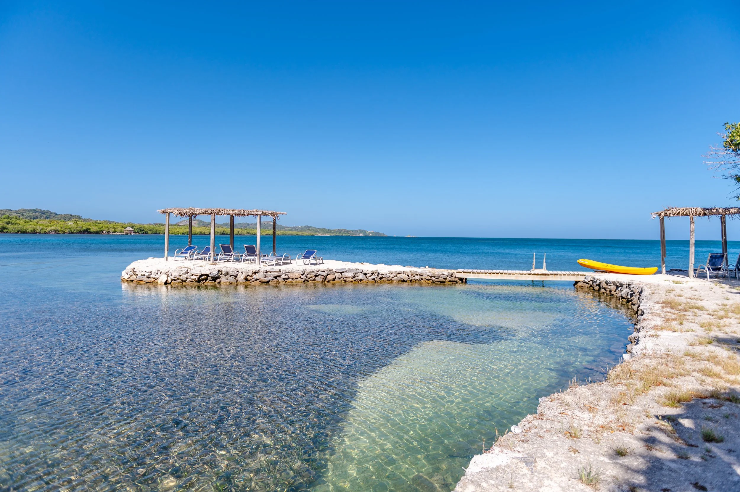 A small stone pier extending into calm, clear blue ocean water with two shaded seating areas on either side and a yellow kayak on the right shore.