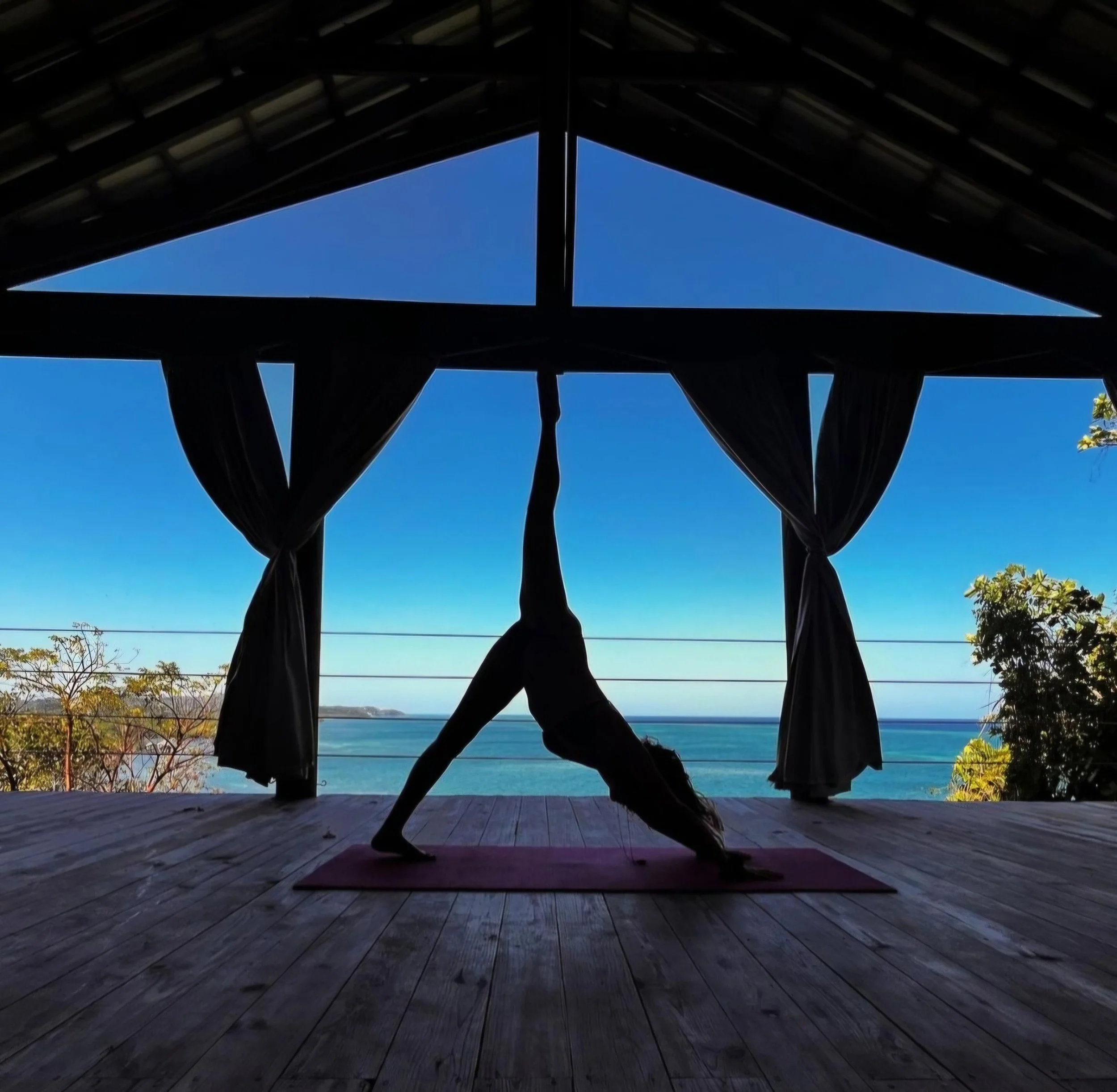 Silhouette of a woman performing a yoga pose on a mat on a wooden deck, overlooking the ocean with blue sky and trees in the background.