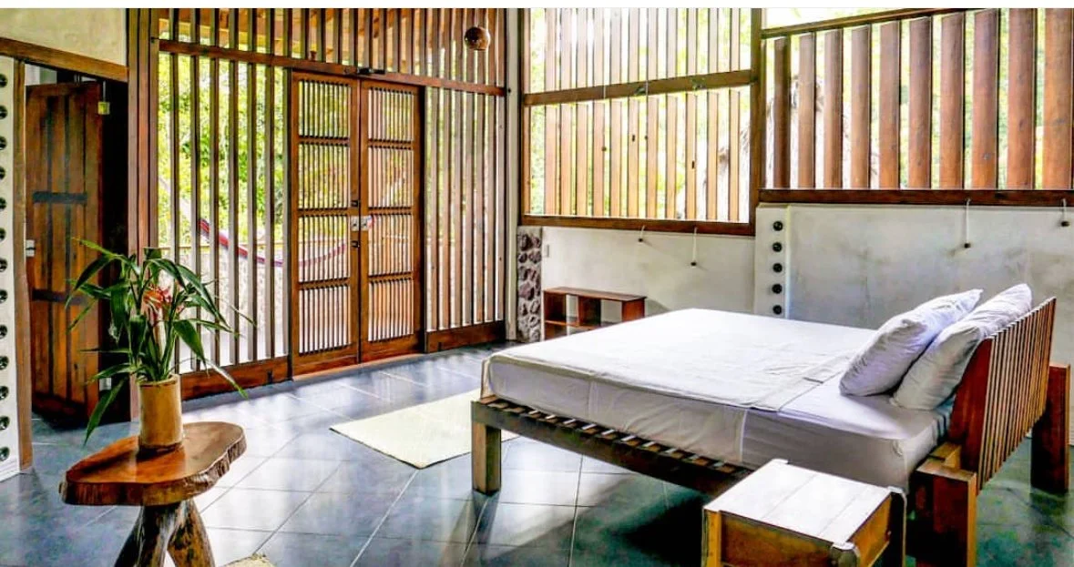 A bedroom with wooden slatted walls, a white bed with pillows, a small side table, a wooden bench, and a plant on a wooden table near a sliding door with wooden lattice design.