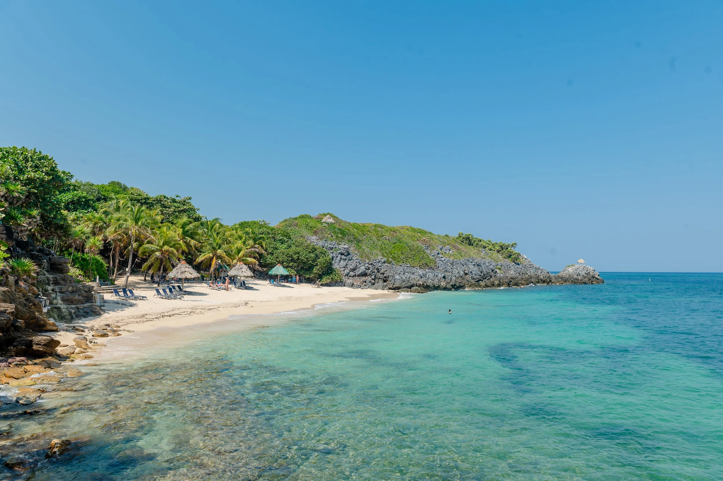 Tropical beach with white sand, palm trees, thatched umbrellas, turquoise water, and rocky hills under a clear blue sky.