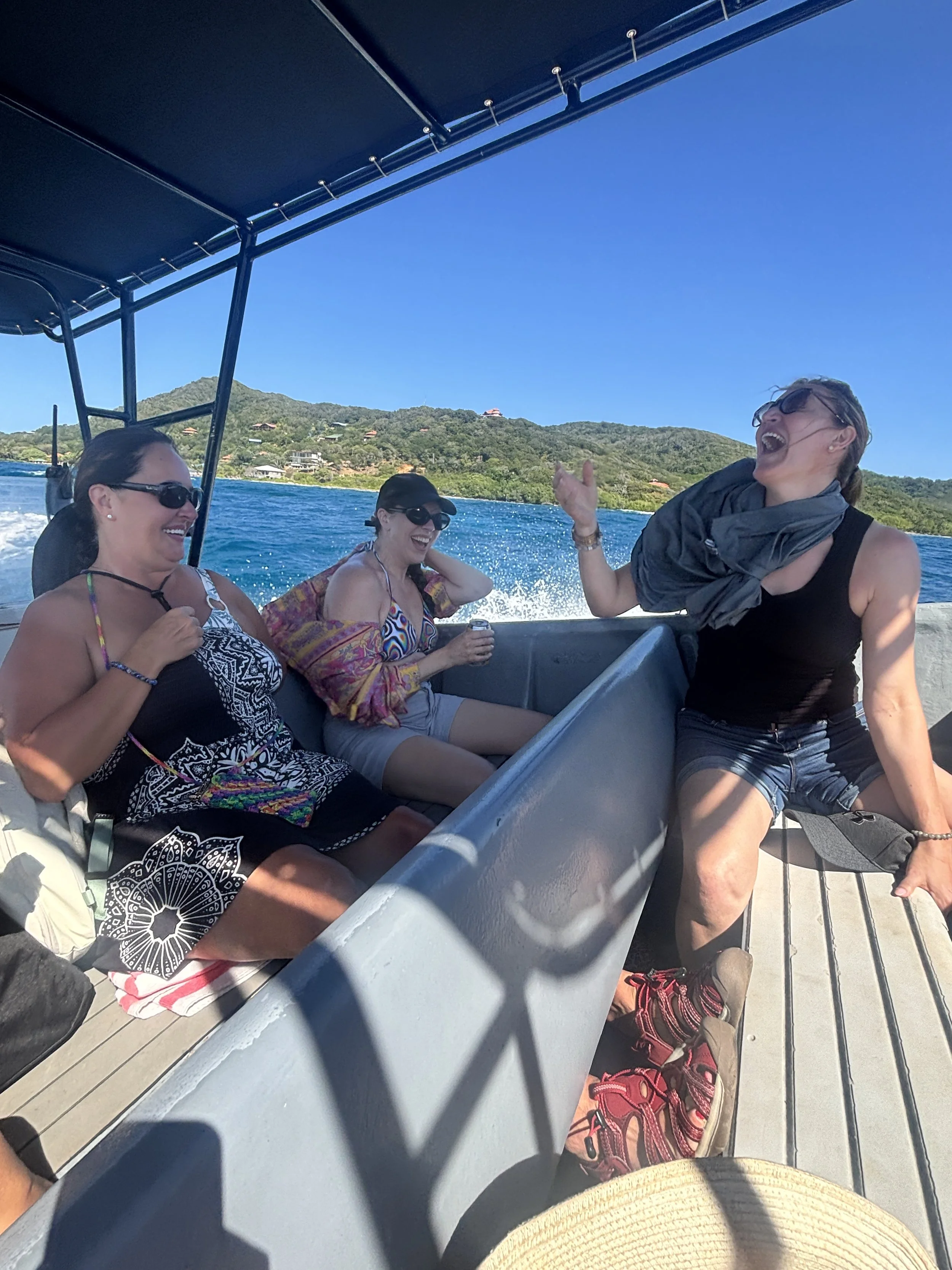 Four women on a boat, laughing and enjoying themselves with a scenic water and mountain background.