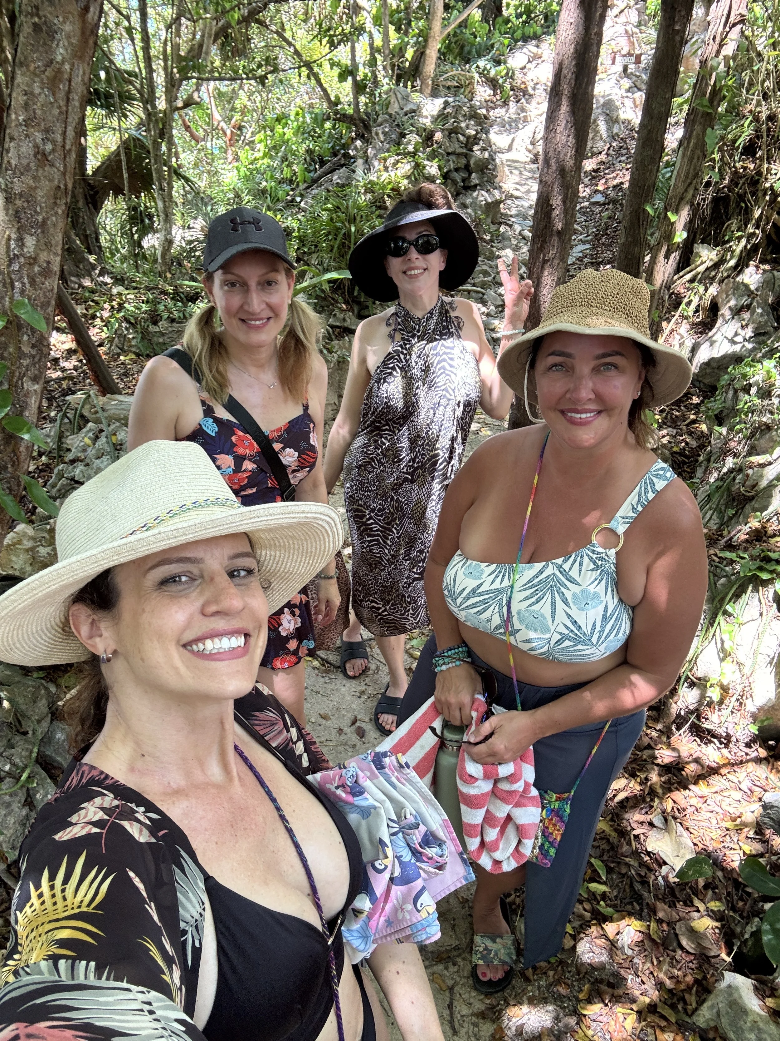 Four women in a forest taking a selfie, all wearing hats and summer clothing.