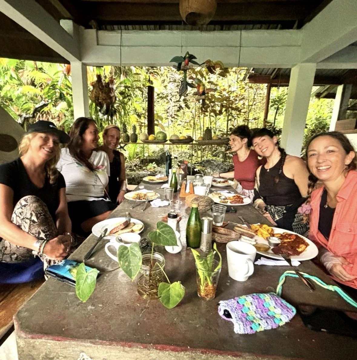 A group of six women sitting around a wooden table outdoors, enjoying a meal in a lush tropical setting with plants and hanging decorations.