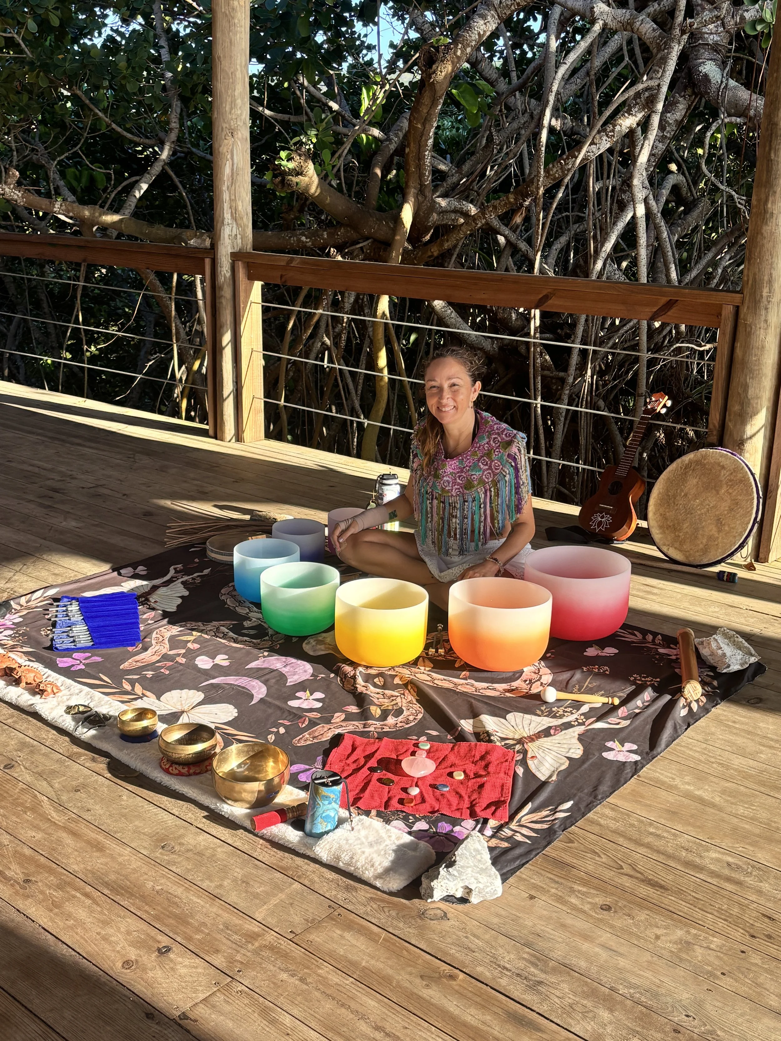 A woman sitting cross-legged on a floral blanket on wooden decking, surrounded by colorful crystal singing bowls, a red cloth with coins and a pink glass orb, and various other musical instruments and stones. She is smiling, with a guitar and a drum 