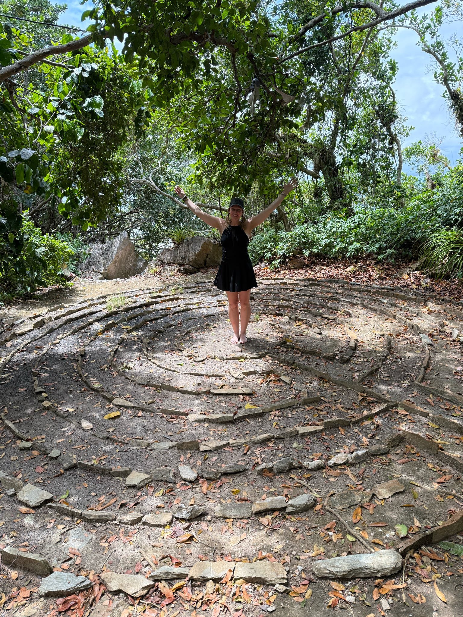 A woman in a black dress and cap standing barefoot on a spiral stone labyrinth in a lush green forest with her arms raised and smiling.