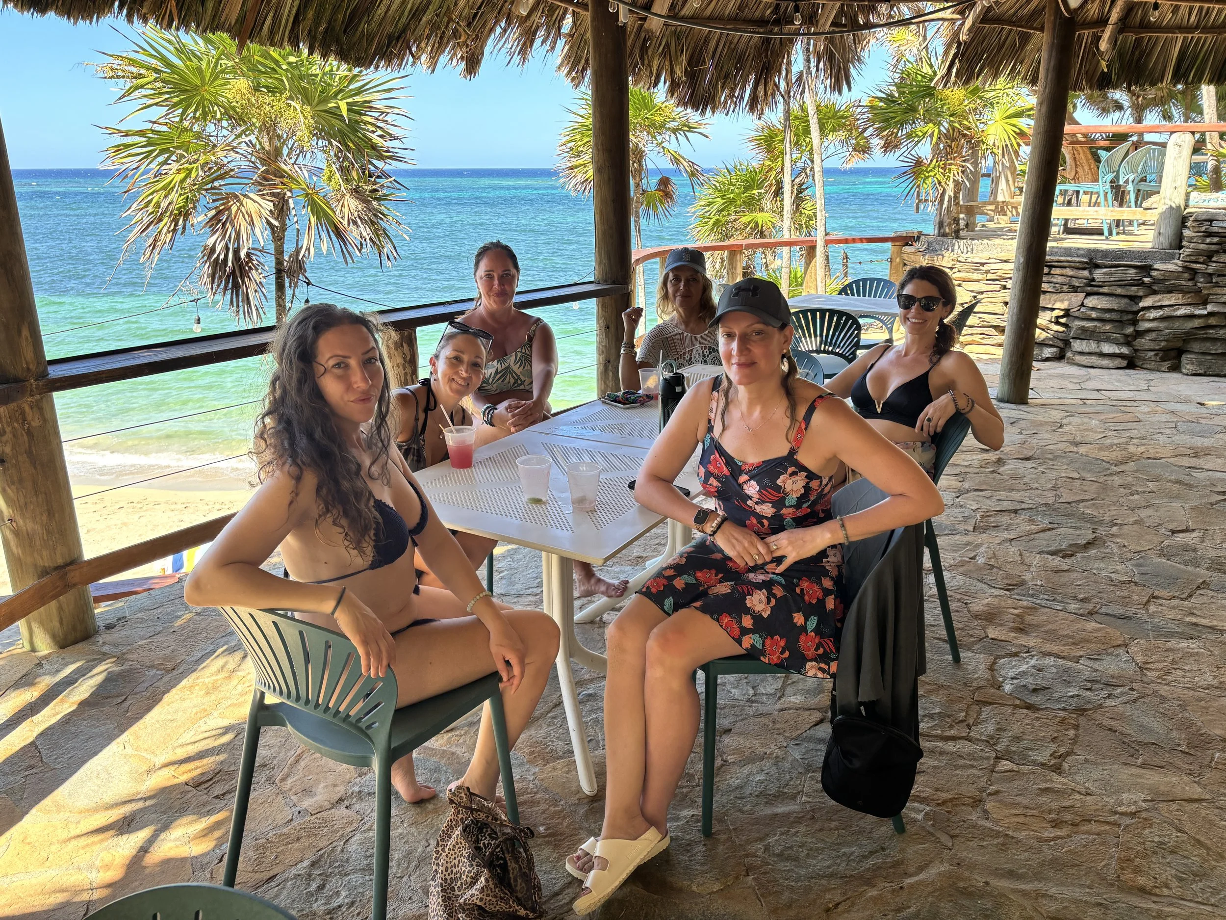 Group of six women sitting at a table on a beachside patio with a view of the ocean and palm trees in the background. They are wearing casual summer clothing and sunglasses, with drinks on the table, enjoying a sunny day.