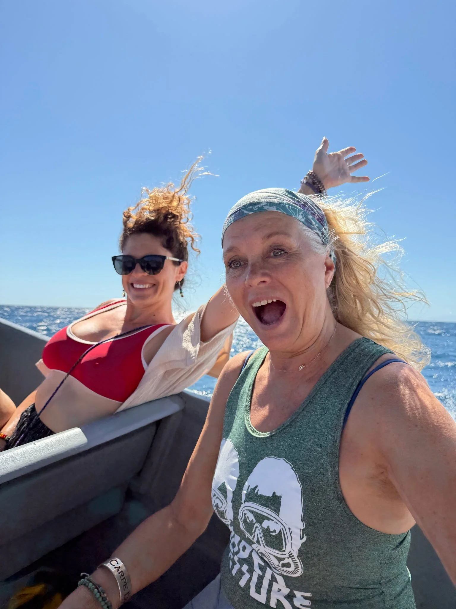 Two women on a boat, one taking a selfie and the other smiling, with the ocean and clear blue sky in the background.