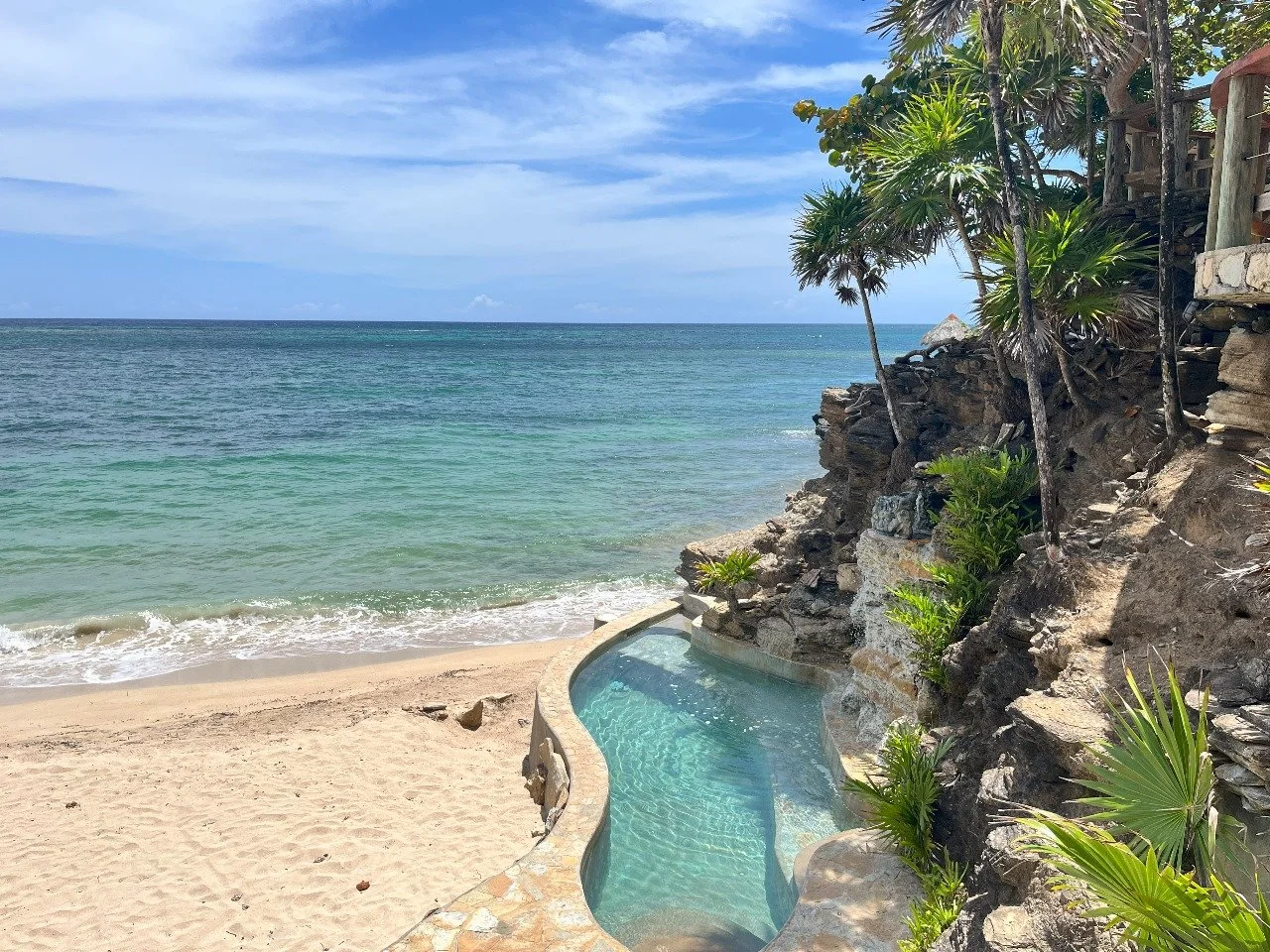 A tropical beach scene with clear blue water, sandy shore, and a small pool built into rocky terrain surrounded by palm trees and lush greenery, under a partly cloudy sky.
